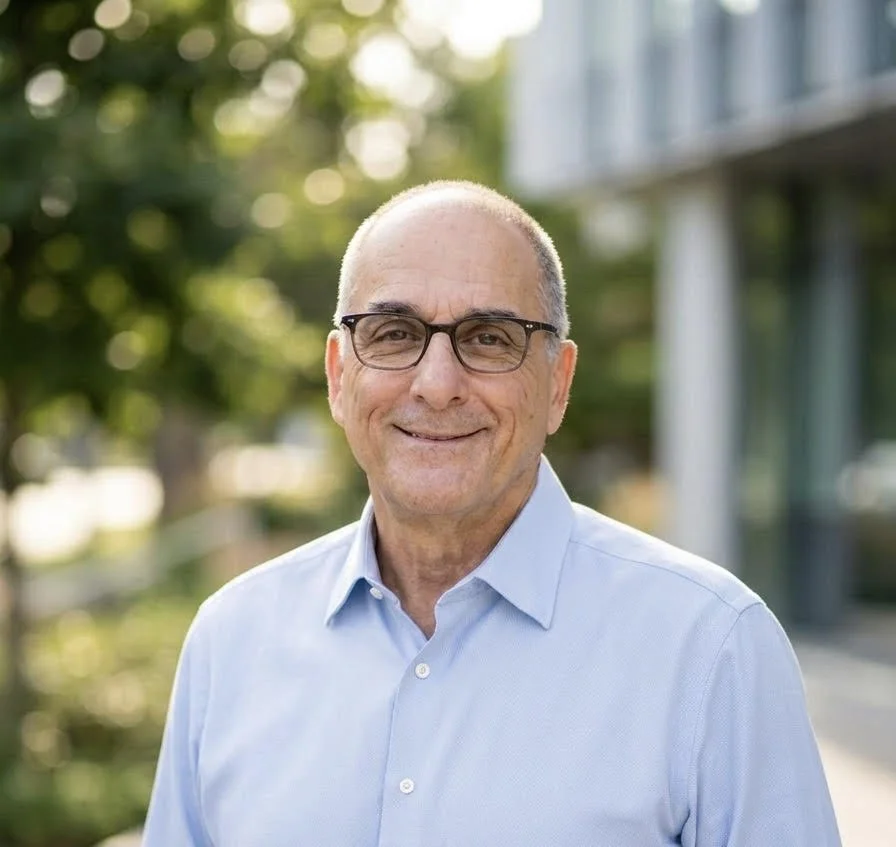 Older man wearing glasses and a light blue button-up shirt smiling outdoors with greenery and a modern building in the background.