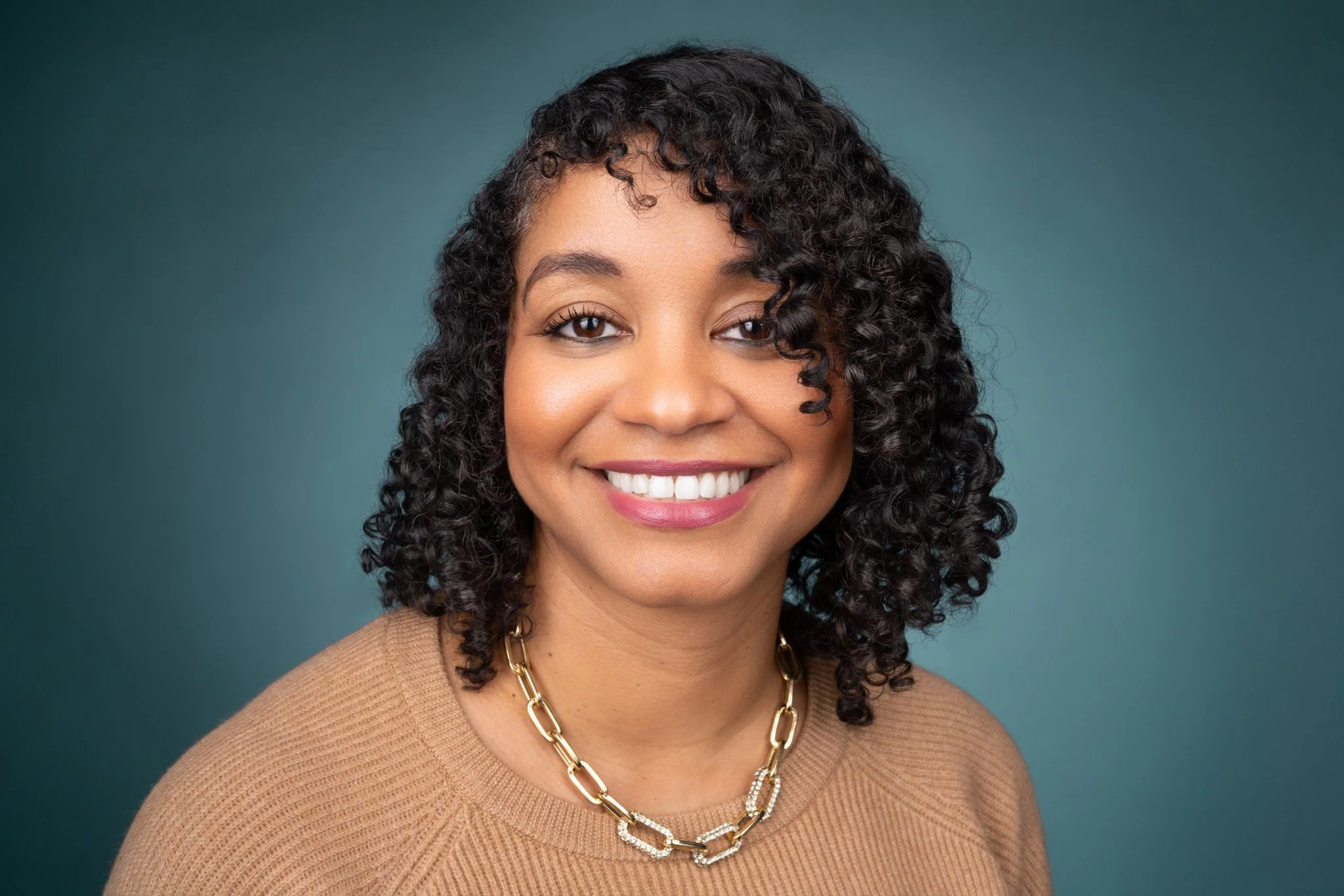 Close-up portrait of a smiling woman with curly dark hair, wearing a beige top and gold necklace, against a teal background.