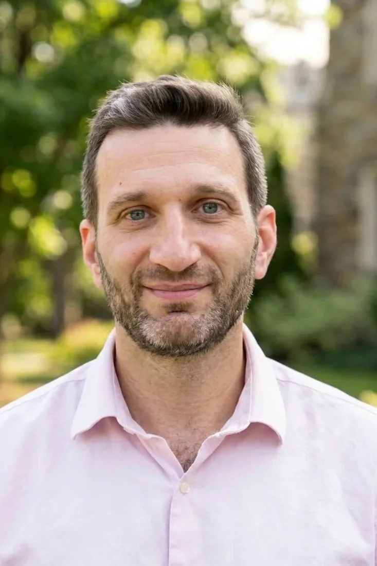A man with light skin, short brown hair, and a beard, smiling and wearing a light pink collared shirt, outdoors with green trees and sunlight in the background.