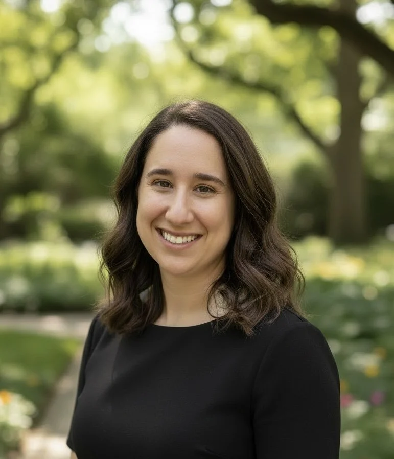 A woman with shoulder-length brown hair smiling outdoors in a park with green trees and flowers in the background.