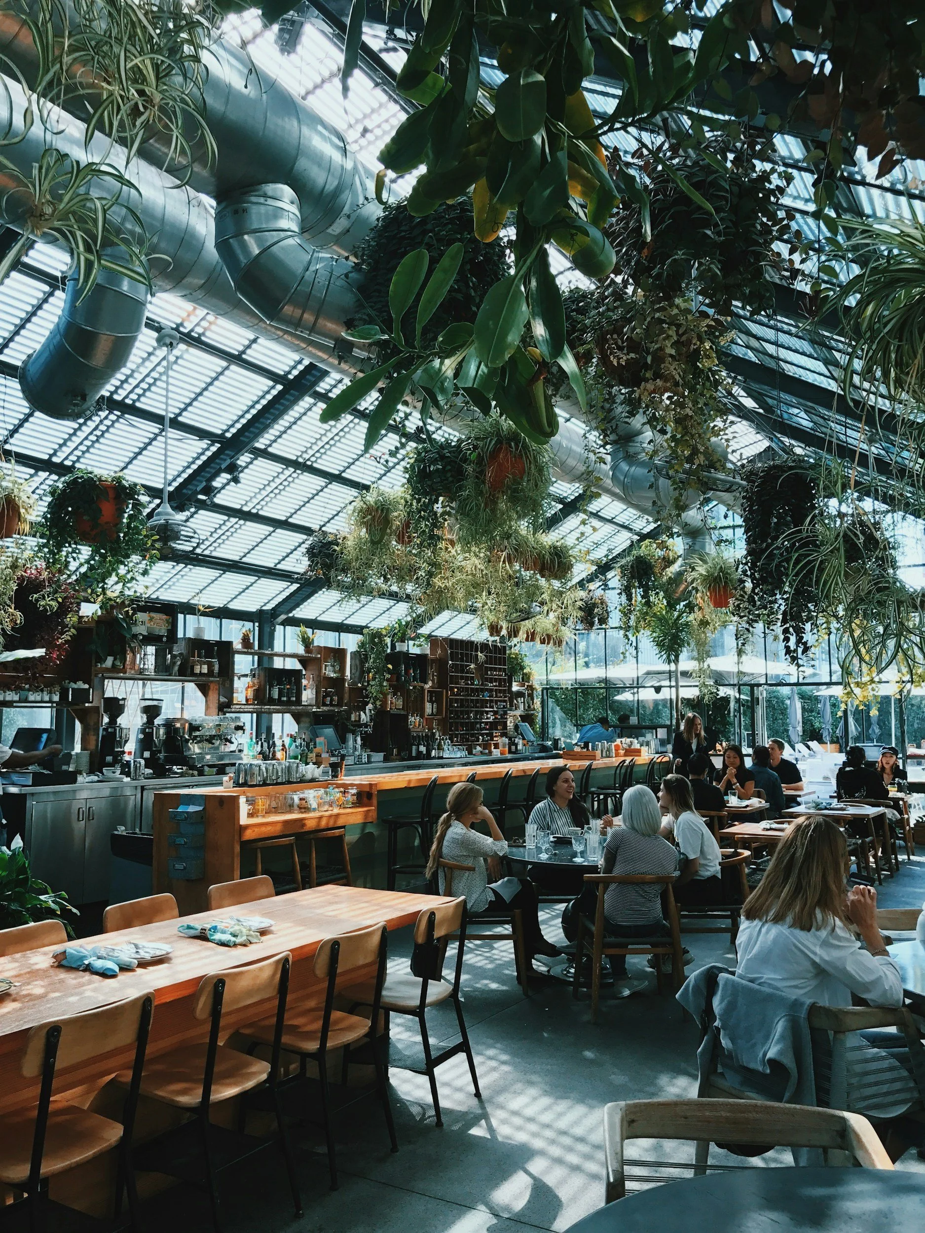 People dining inside a greenhouse-style restaurant with hanging plants and large metallic ventilation pipes overhead.