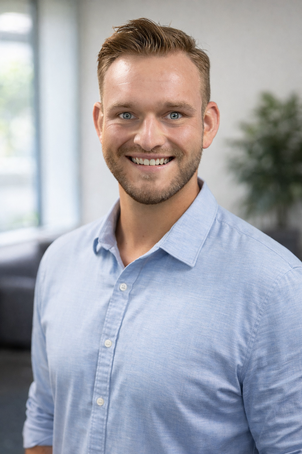 A smiling man with short, styled hair and a beard, wearing a light blue button-up shirt, in an office setting with a window and a plant in the background.