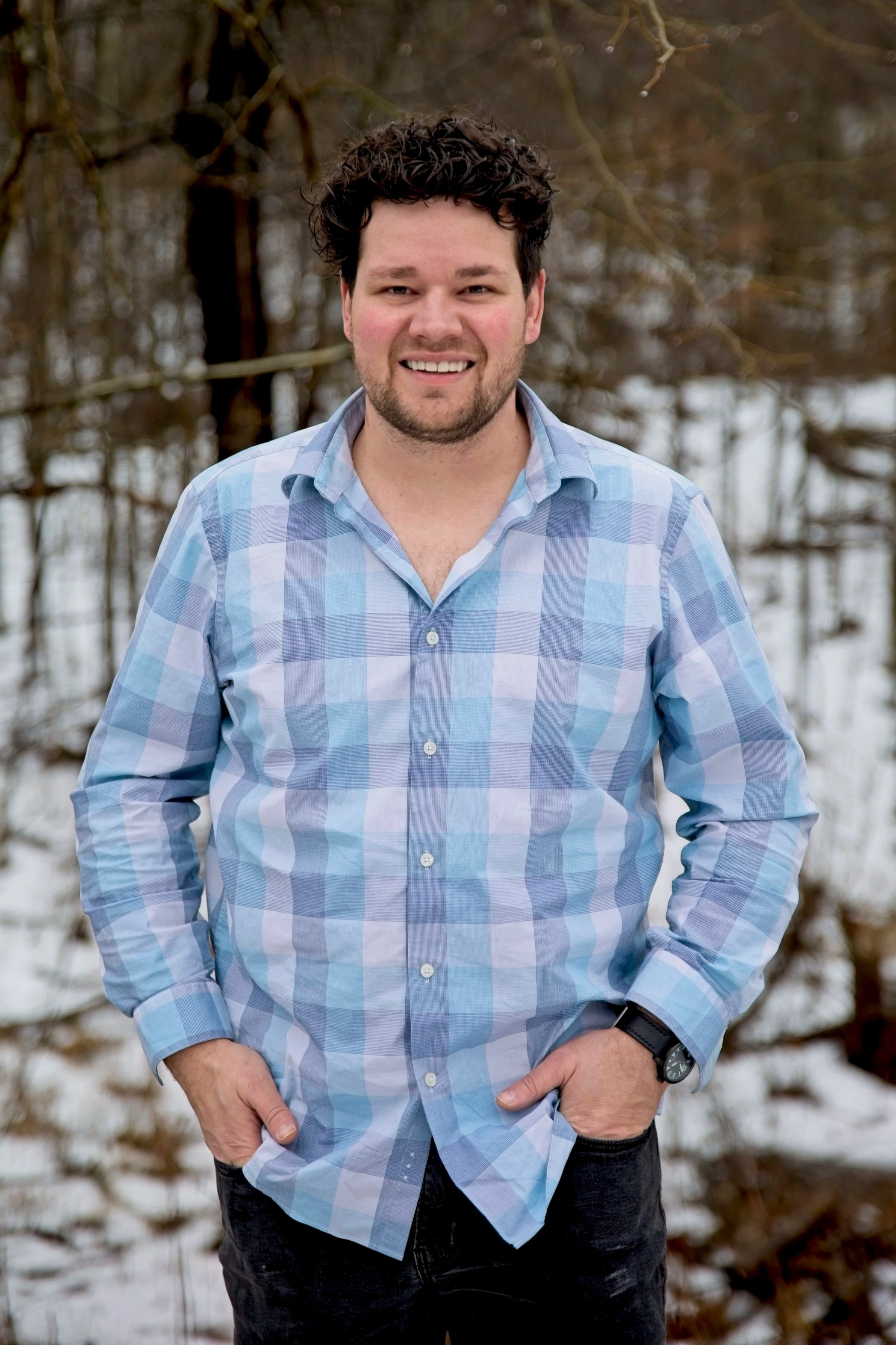 A smiling man with curly dark hair wearing a light blue checkered shirt, standing outdoors in a snowy, wooded area.