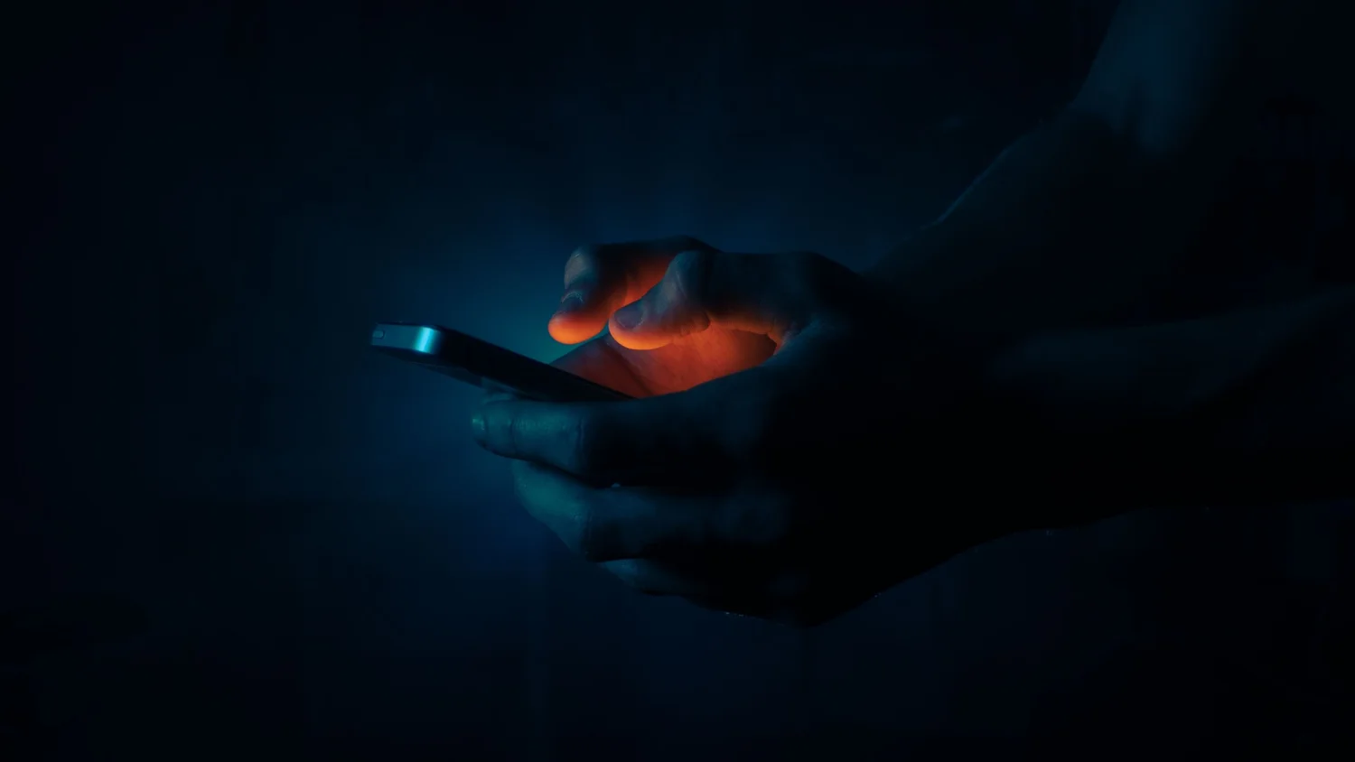 A hand illuminated by a glowing phone screen in a dark room, late at night.