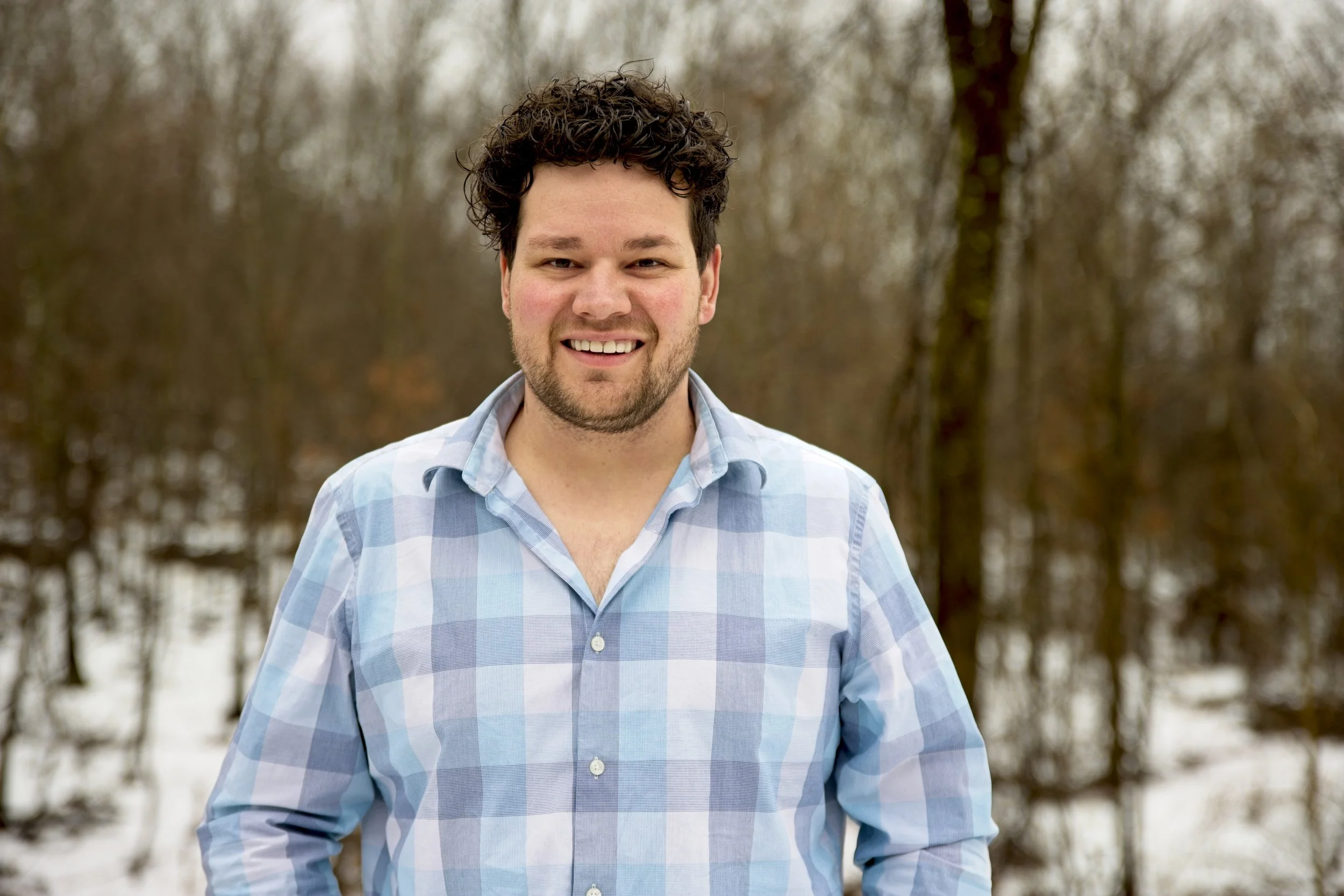 A smiling man with dark curly hair and a beard, wearing a blue and white plaid shirt outdoors in a wintery forested area.