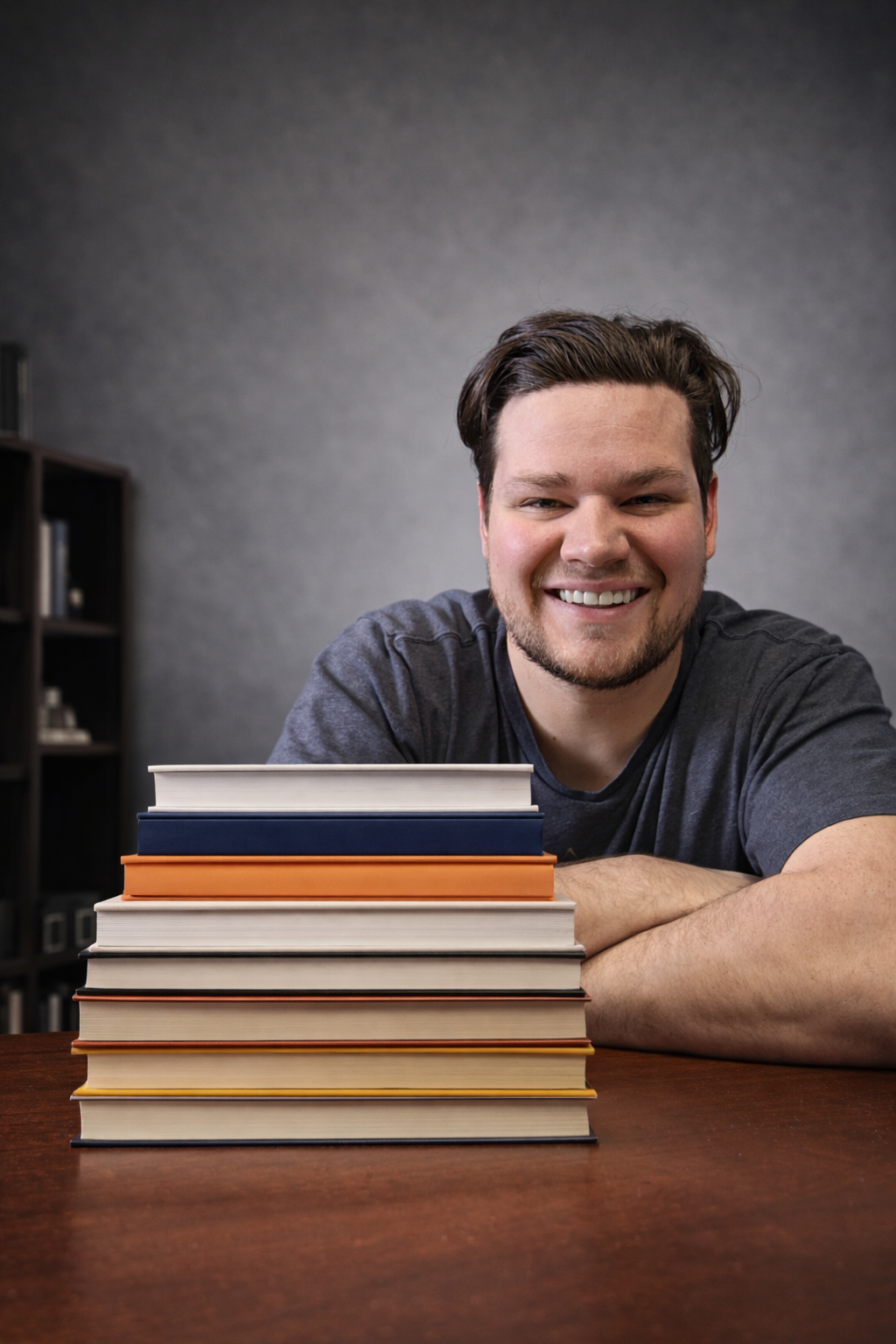 A smiling young man with dark hair and a beard resting his chin on his crossed arms at a table, with a stack of books in front of him and a bookshelf in the background.