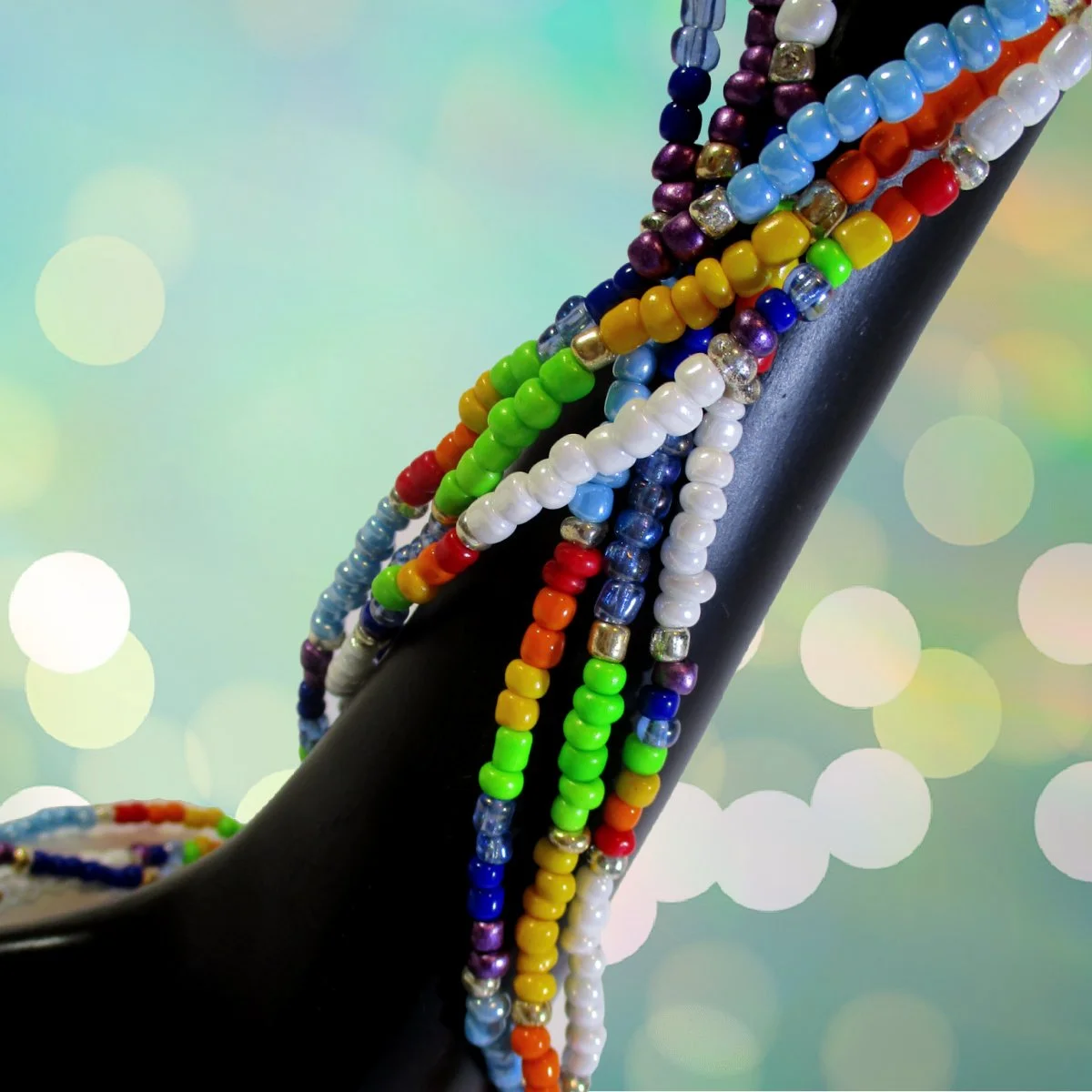Colorful beaded necklaces draped over a black high heel shoe against a soft focus pastel background.