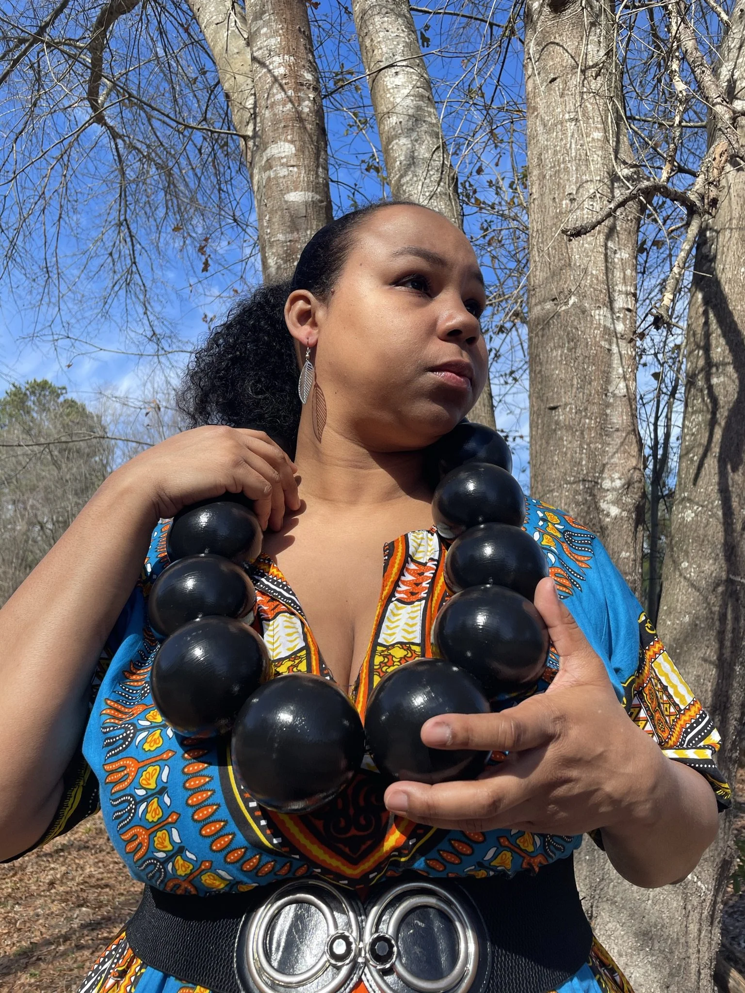 A woman outdoors in a colorful African-patterned dress and large black beaded necklace, standing against a backdrop of trees and a blue sky, looking thoughtfully to the side.