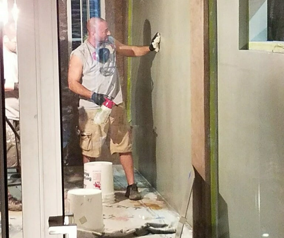 A man working on a construction project indoors, standing on a covered floor with paint buckets around him, holding a caulking gun, and applying a material to the wall.