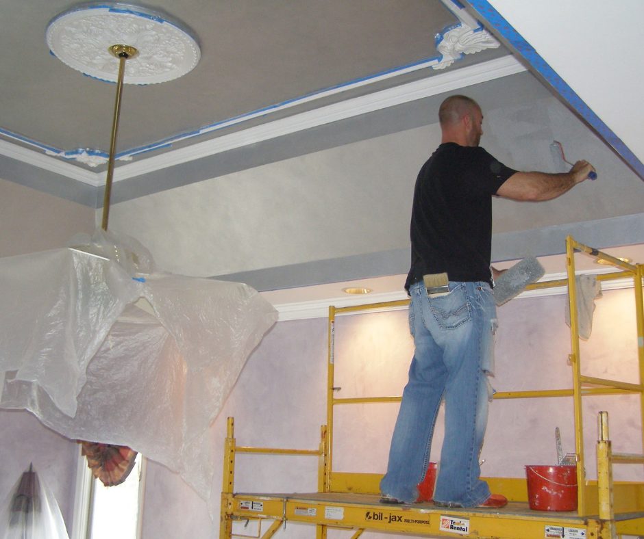 A man is painting the ceiling of a room, standing on a yellow scaffolding. The ceiling has decorative molding, and protective plastic sheeting is hanging from the ceiling and covering parts of the room while he works.