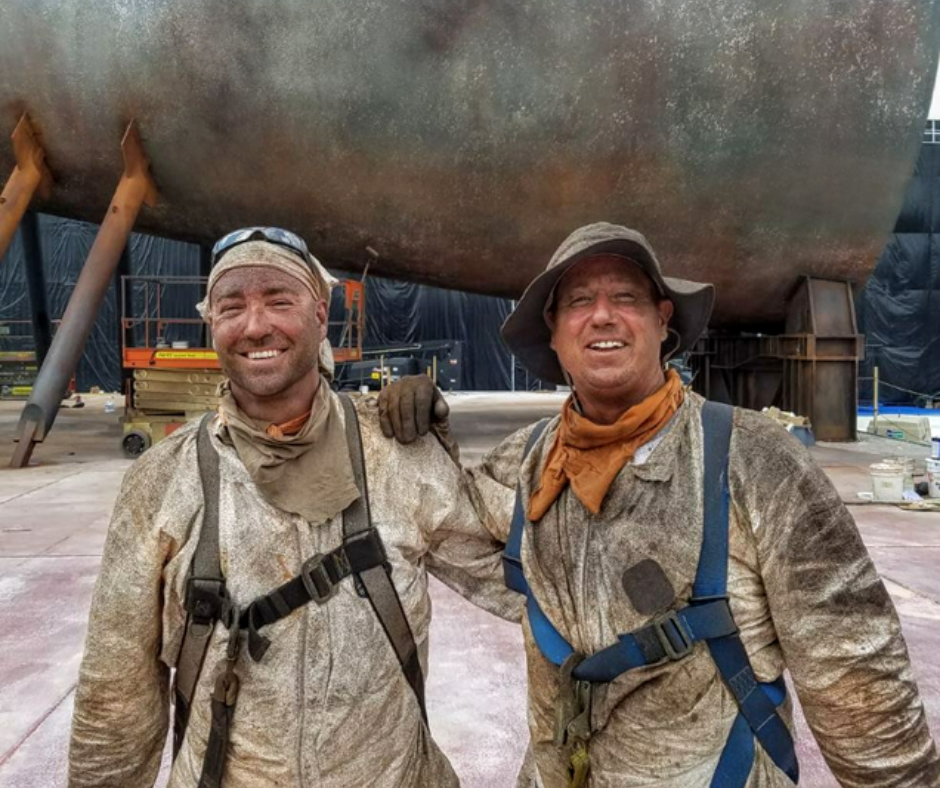 Two smiling workers in protective gear and dust masks, standing in front of a large ship hull in a shipyard or dry dock.