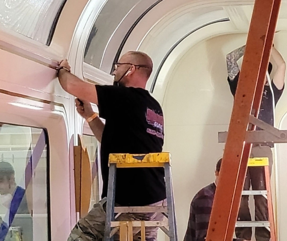 A man working on a window frame inside a building, standing on a yellow step ladder, with another orange ladder nearby. Another person is in the background, and a reflective glass surface shows people sitting at tables.