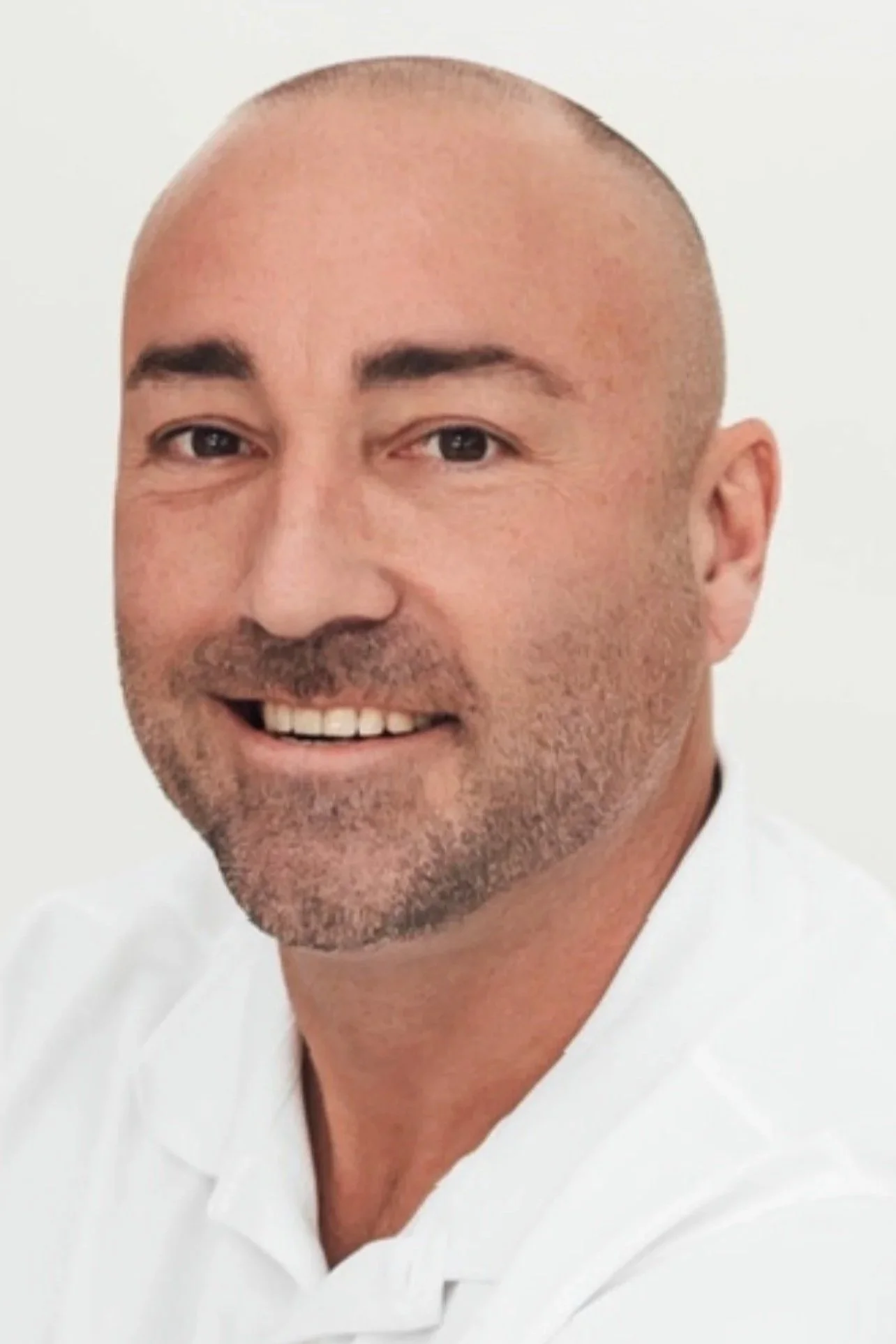 Close-up of a smiling man with a shaved head, dark eyebrows, and stubble, wearing a white shirt against a white background.