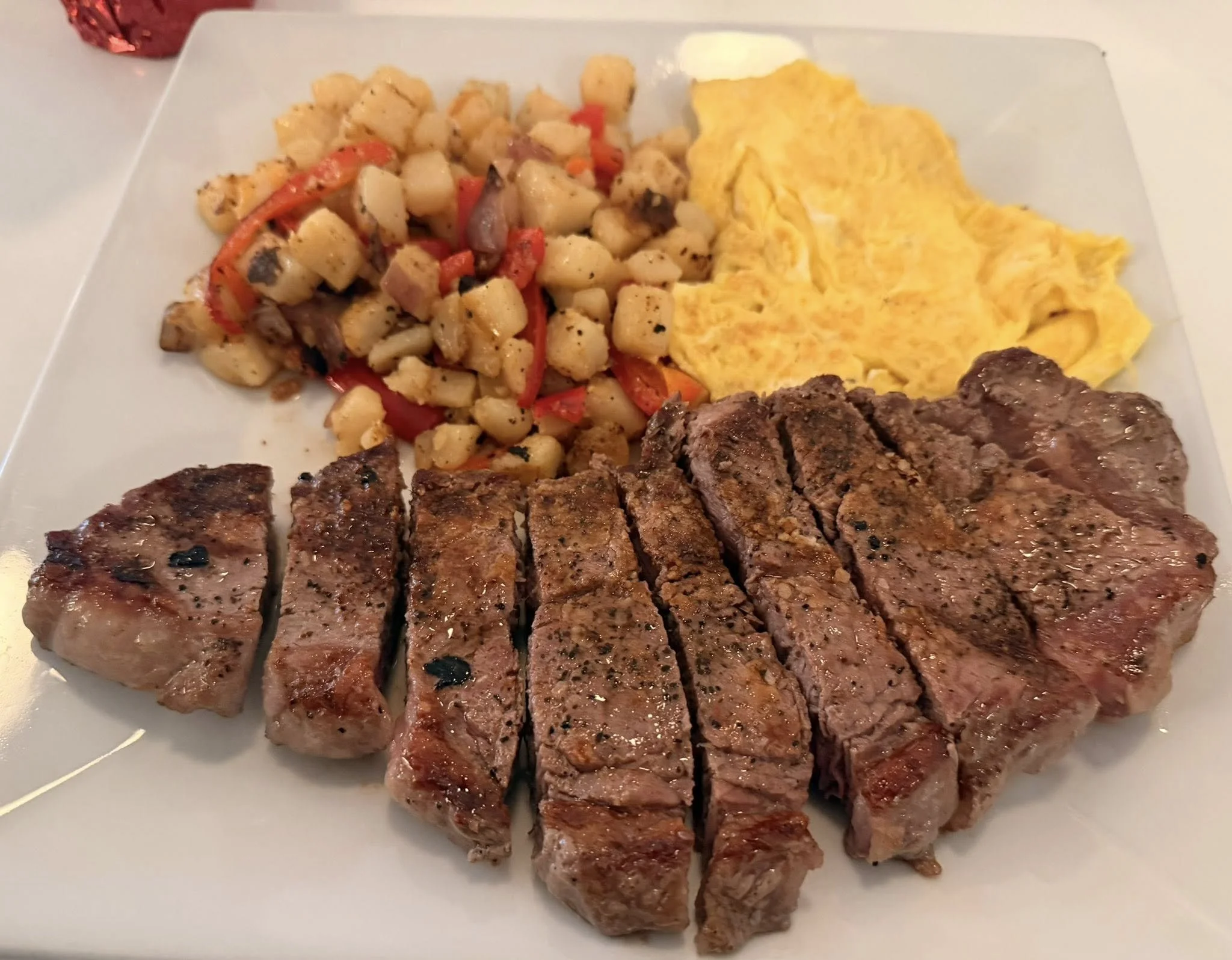 A plate with sliced grilled steak, scrambled eggs, and a hash mixed with potatoes, red peppers, and mushrooms.
