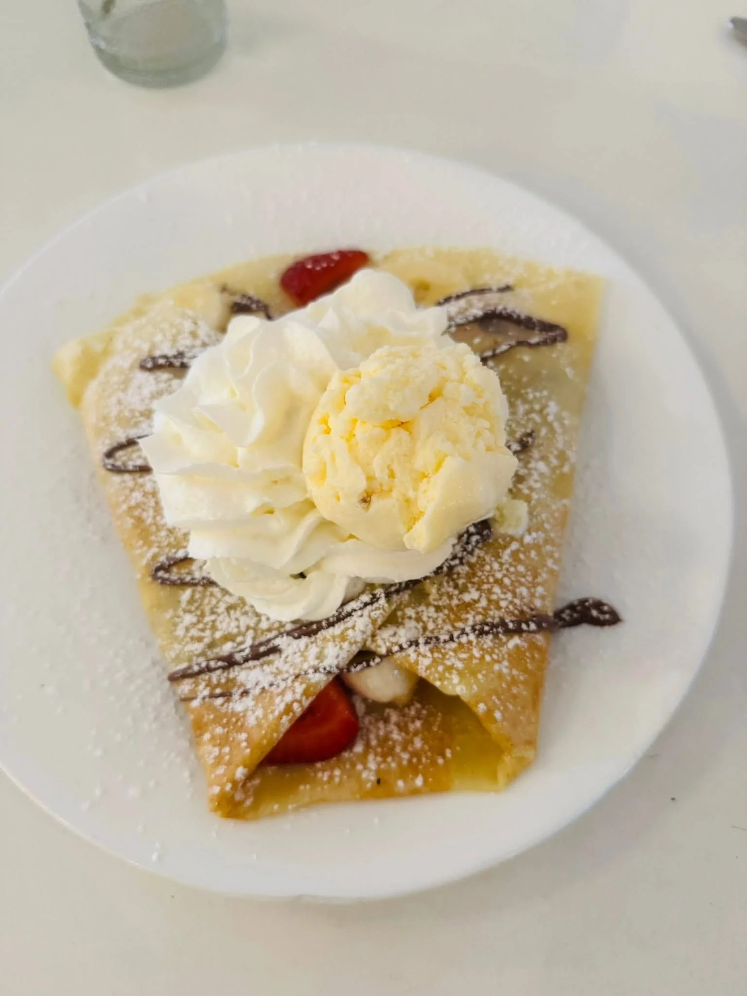 A dessert crepe on a white plate topped with whipped cream, vanilla ice cream, chocolate drizzle, and powdered sugar, with strawberries inside.