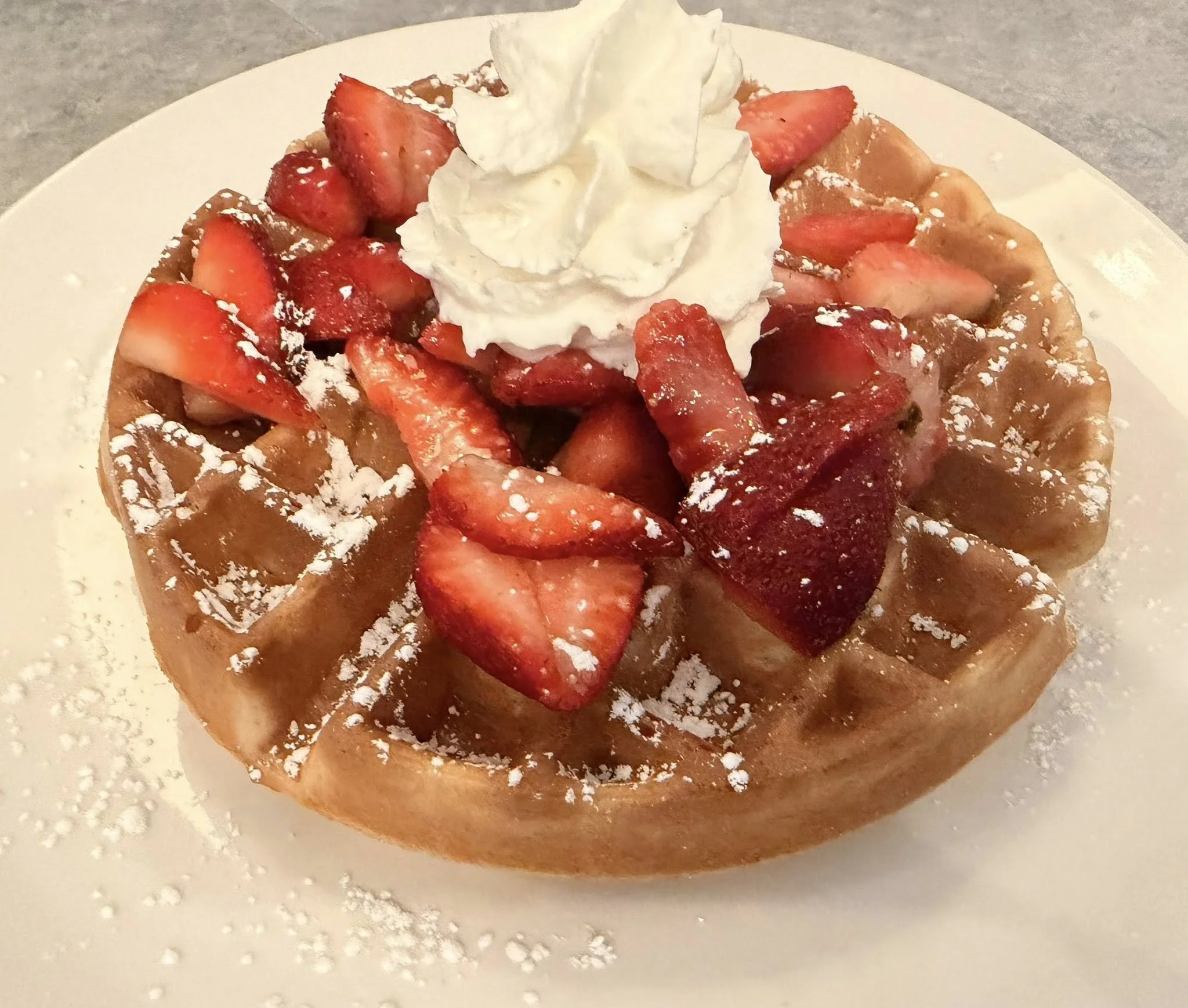 Waffle topped with sliced strawberries, whipped cream, and powdered sugar, served on a white plate.