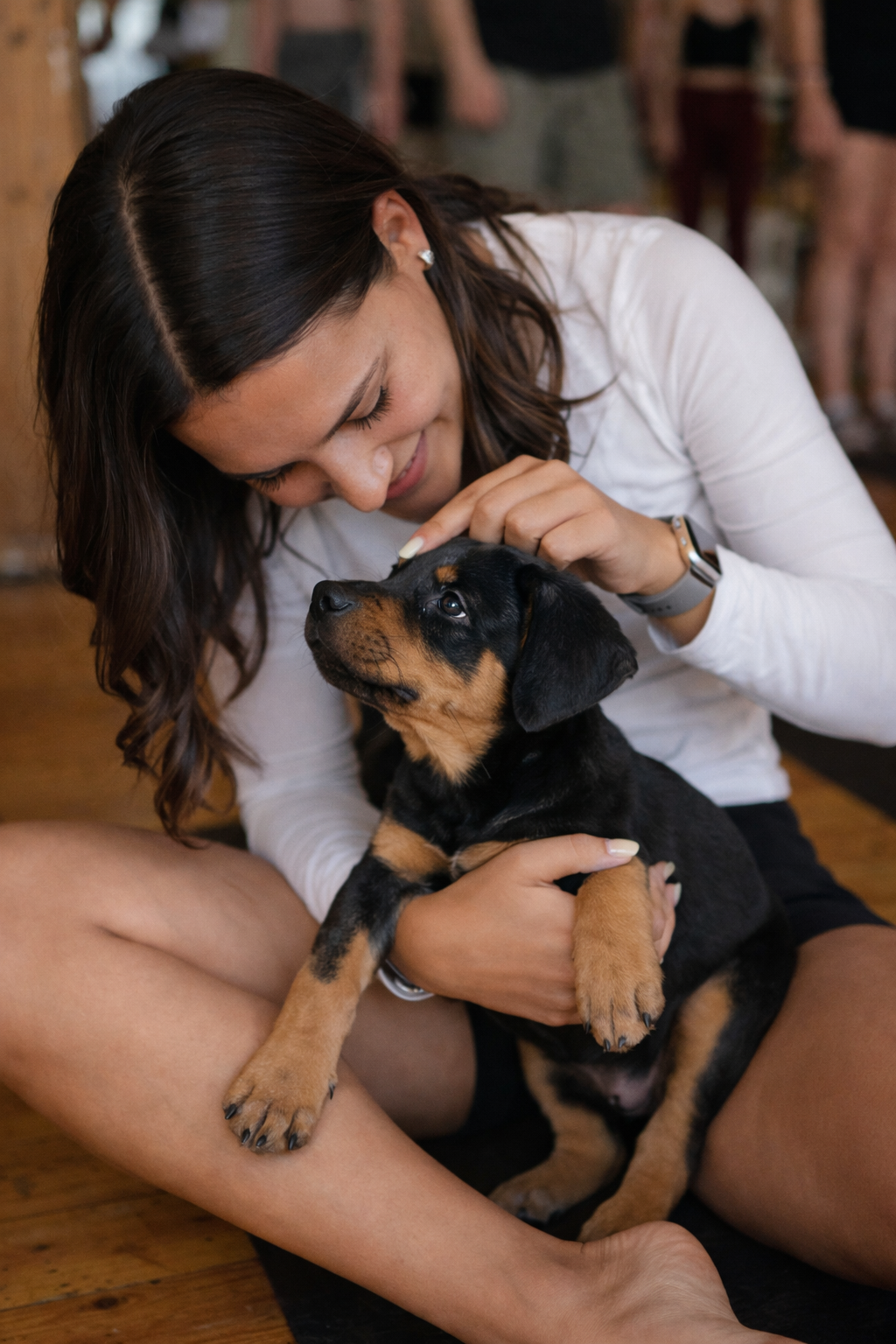 A woman smiling and gently holding a cute black and tan puppy on her lap, sitting on a wooden floor indoors.