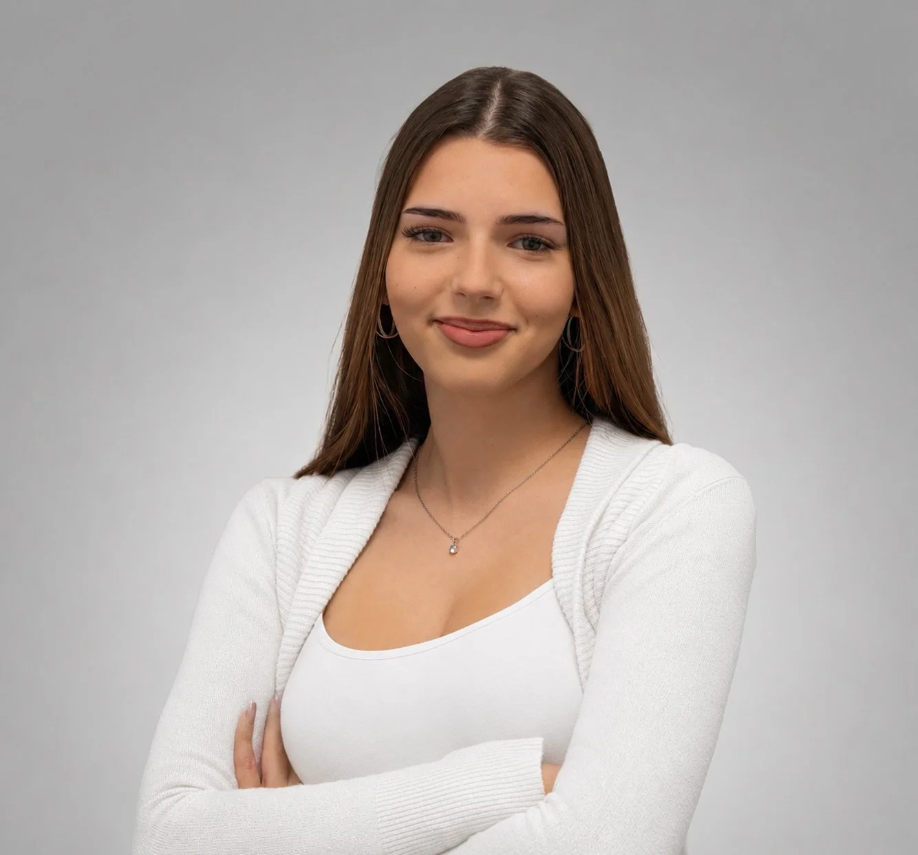 Junge Frau mit braunen Haaren, weißem Top und weißem Pullover, lächelnd, vor grauem Hintergrund.