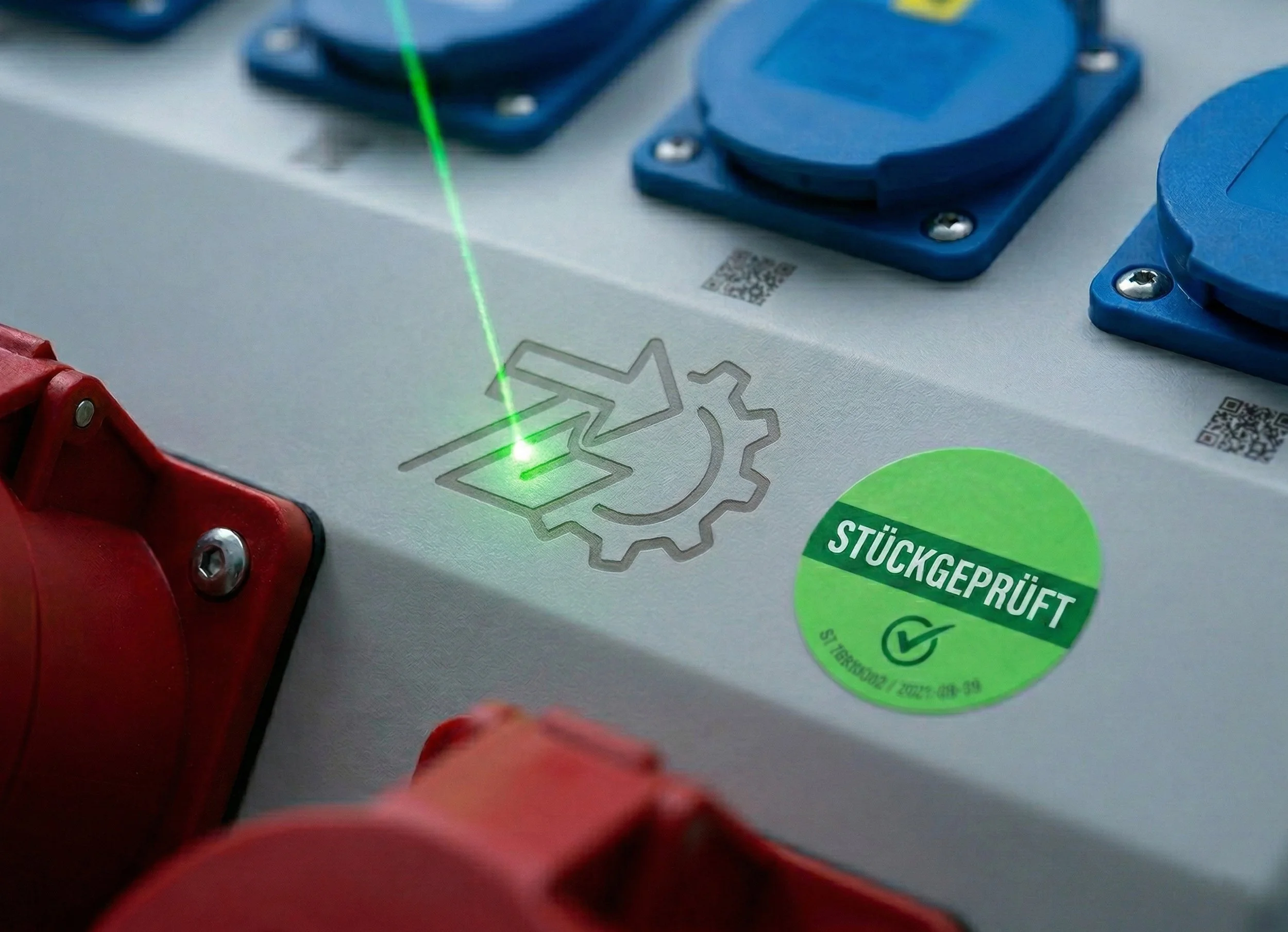Grüner Laserstrahl trifft auf eine Oberfläche mit Aufklebern, darunter ein grüner Aufkleber mit der Aufschrift 'STÜCKGEPRÜFT' und QR-Codes.