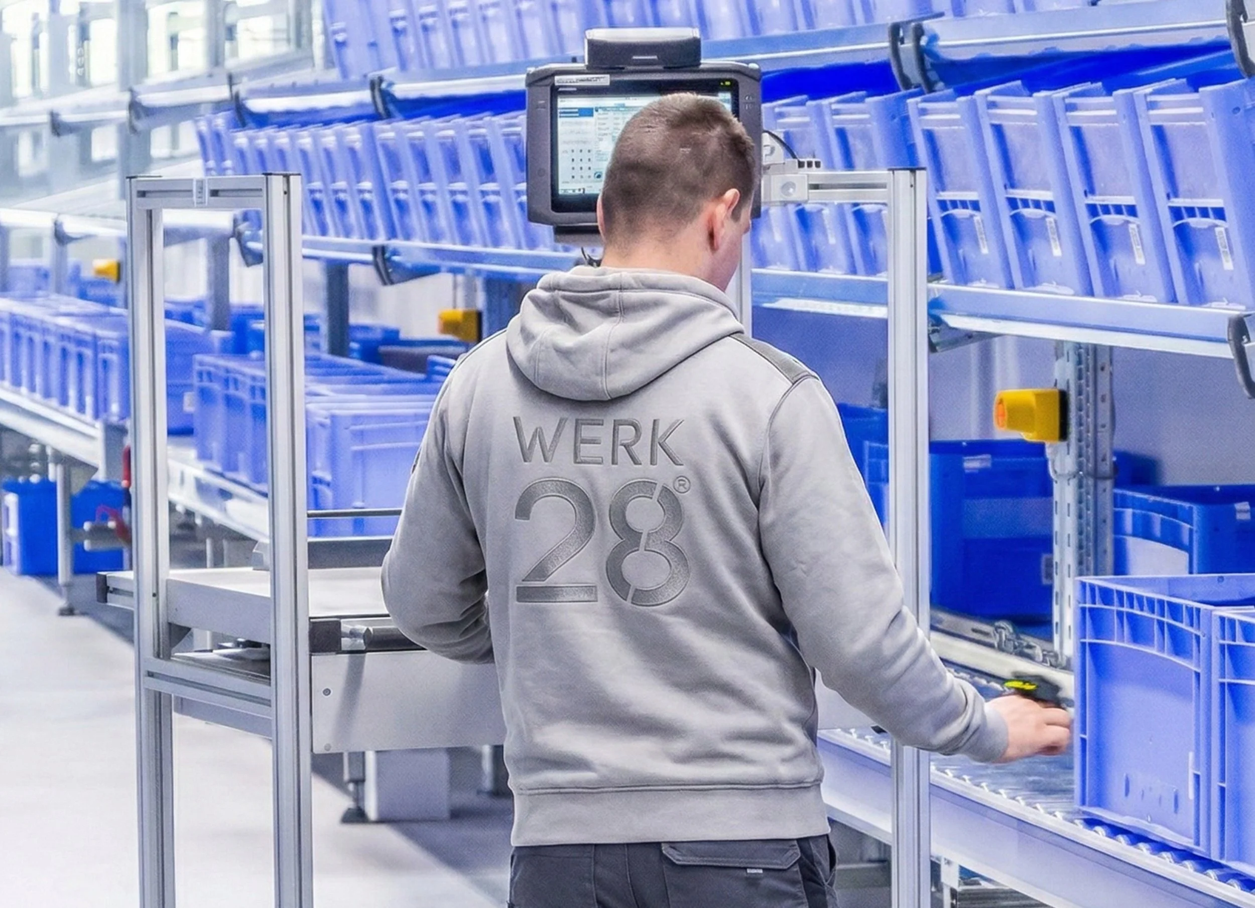 Ein Mann arbeitet in einem Lager oder einer Fabrik, umgeben von blauen Lagerregalen und einem Computer. Er trägt einen grauen Hoodie mit der Aufschrift 'WERK' auf dem Rücken.