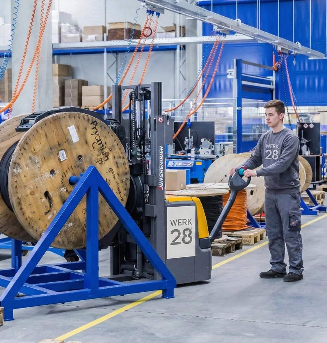Ein junger Mann bedient einen elektrischen Steuerschlauch in einer Fabrikhalle, die mit großen Spulen aus Draht, Werkzeugen und Lagerregalen gefüllt ist.