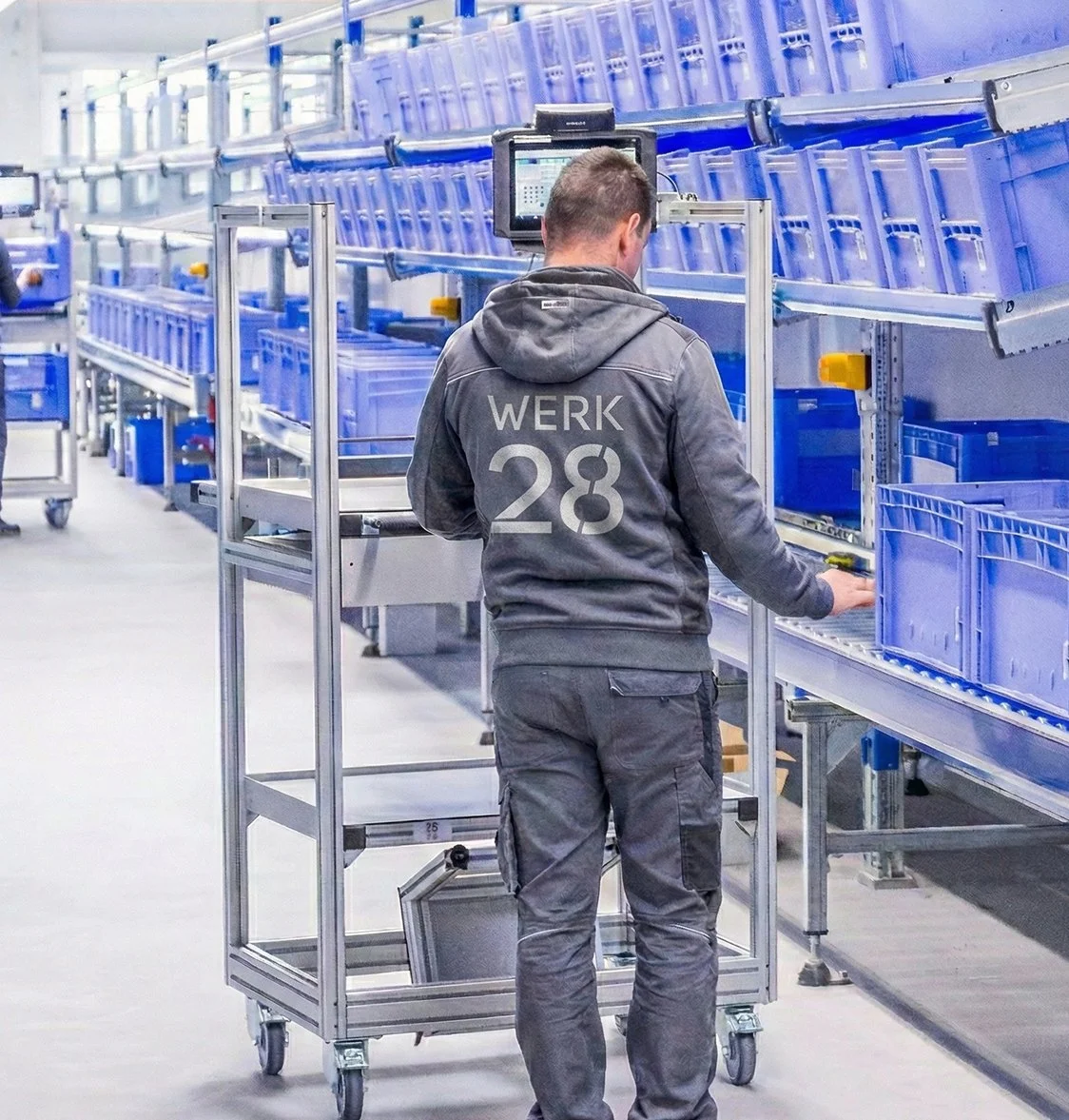 Mann in Arbeitskleidung mit der Aufschrift 'WERK 28' arbeitet in einer Fabrikhalle mit blauen Lagerkisten auf metallenen Eisenregalen.