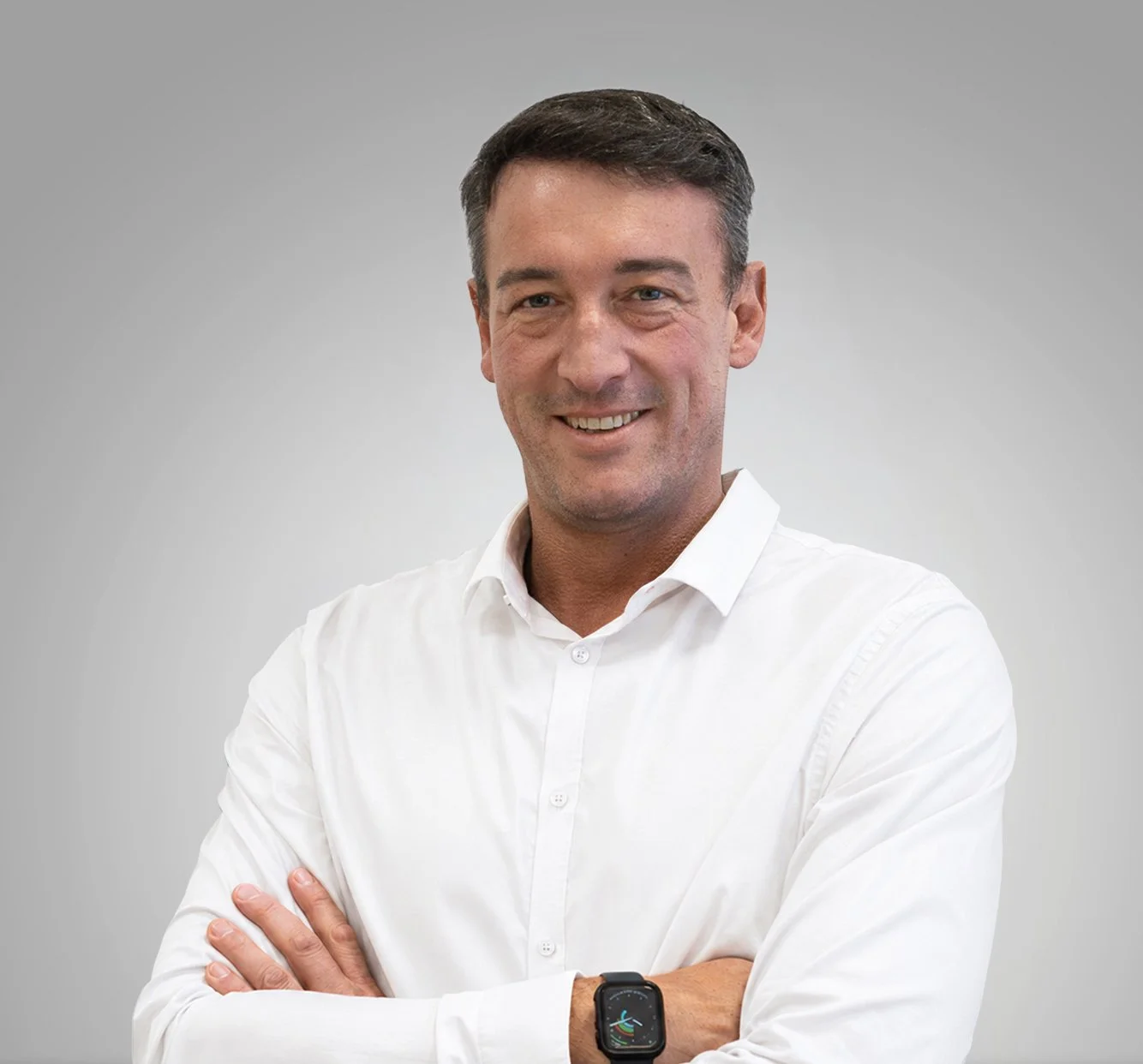 Porträt eines Mannes in weißem Hemd, lächelnd, mit verschränkten Armen, trägt eine Smartwatch, vor einfarbigem grauem Hintergrund.