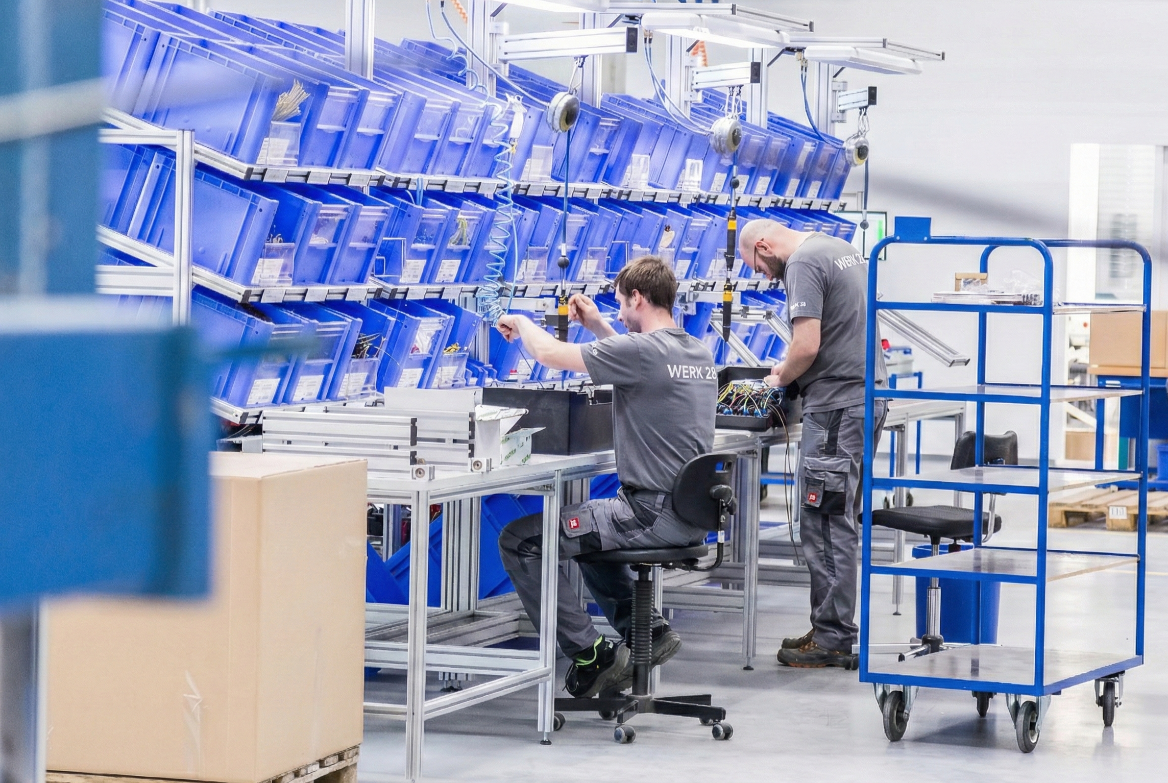 Zwei Männer arbeiten in einer Fertigungs- oder Montagshalle mit blauen Aufbewahrungsboxen auf Regalen und Werkbänken.