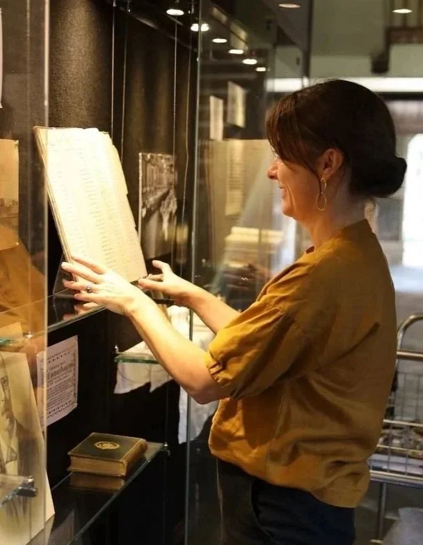 A woman reading a book in a glass display case at a museum or library.