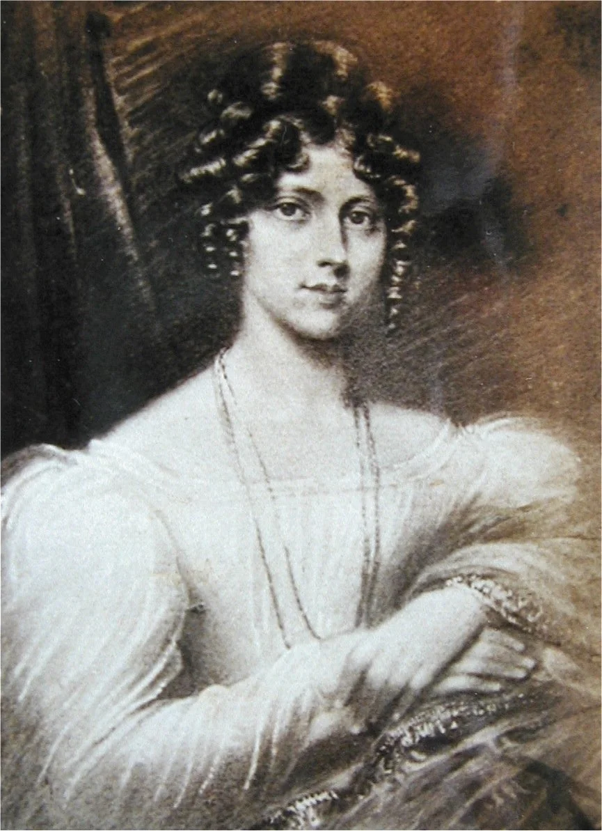 A black-and-white portrait of a woman with curly hair, wearing a light-colored dress and a necklace, sitting with her arms crossed on a surface.