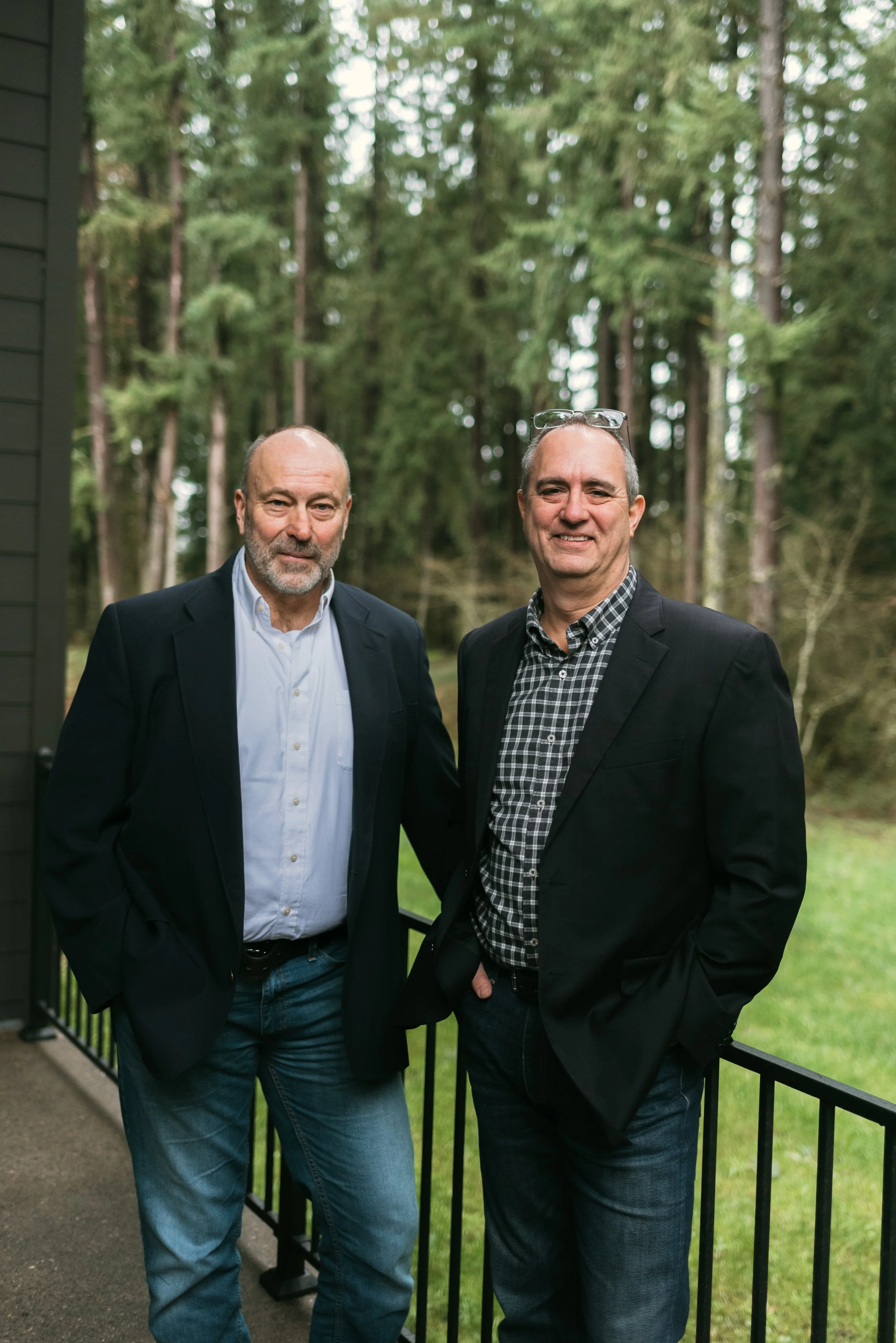 Two middle-aged men in suits standing outdoors on a porch with a forest background, smiling at the camera.