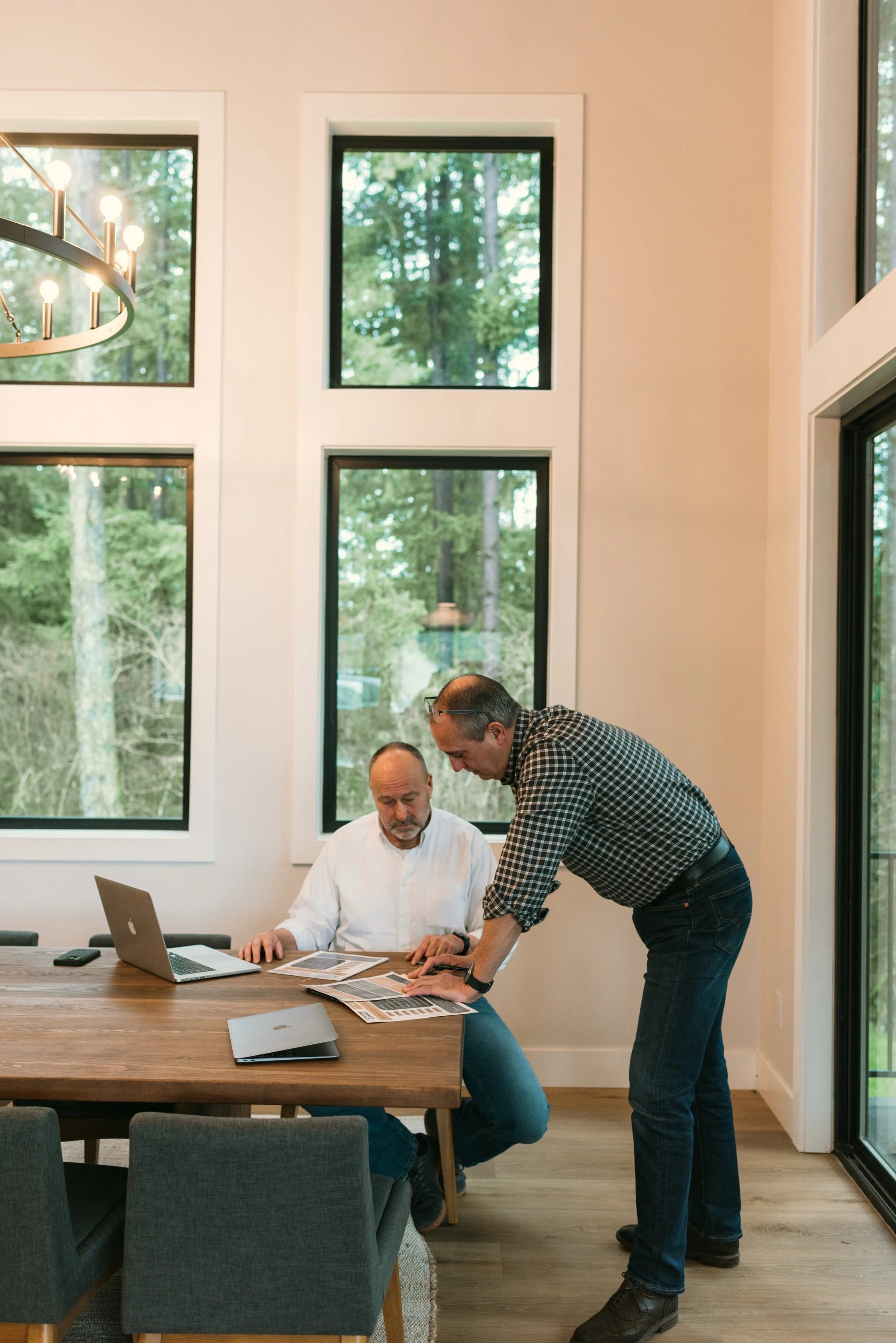 Two men in a meeting room looking at documents on a table. One is seated, working on a laptop, while the other is standing, leaning over to see the documents, with large windows showing trees outside behind them.