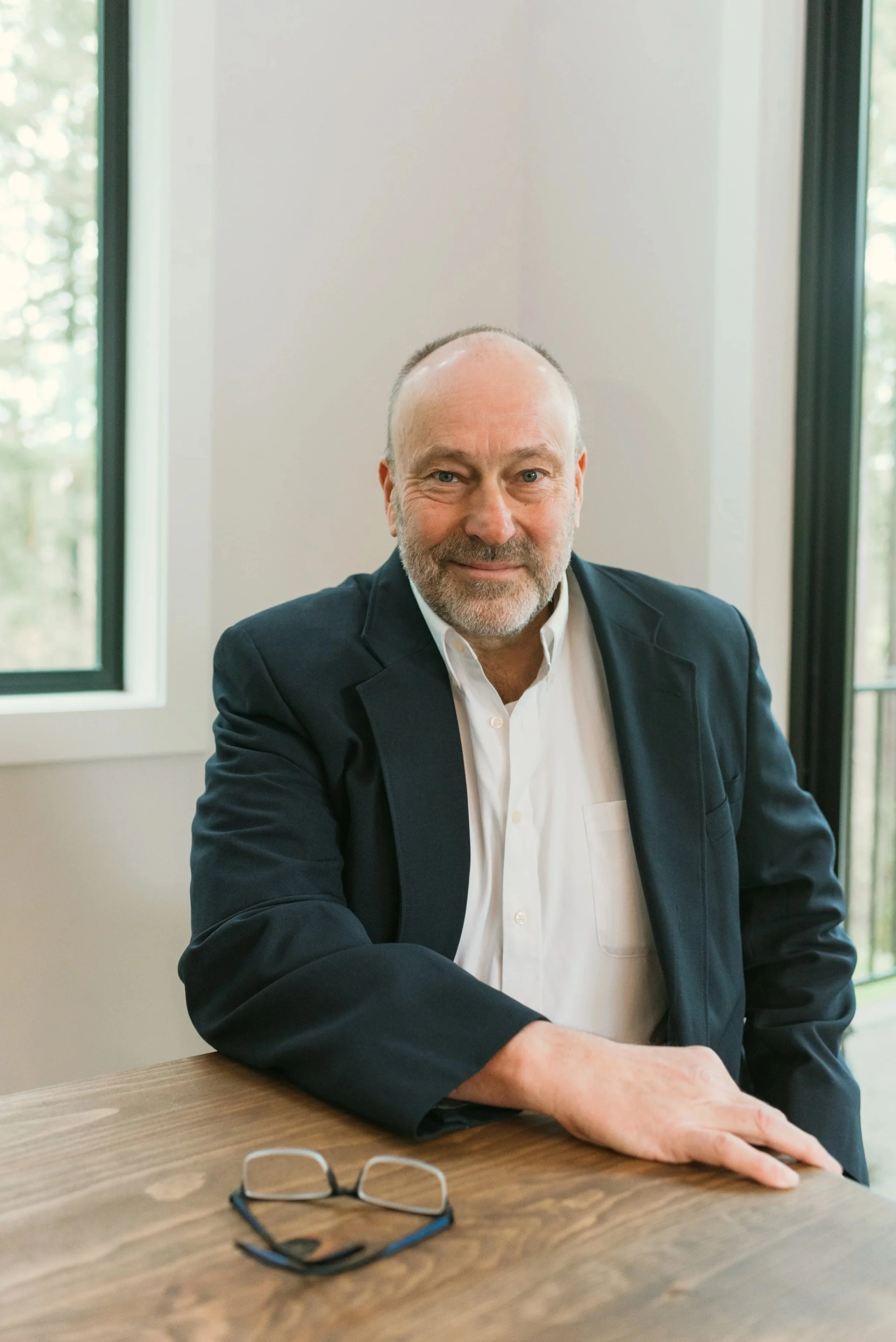 A middle-aged man with a beard in a dark suit and white shirt is sitting at a wooden table, smiling slightly. A pair of eyeglasses rests on the table in front of him, and he is near a window with greenery visible outside.