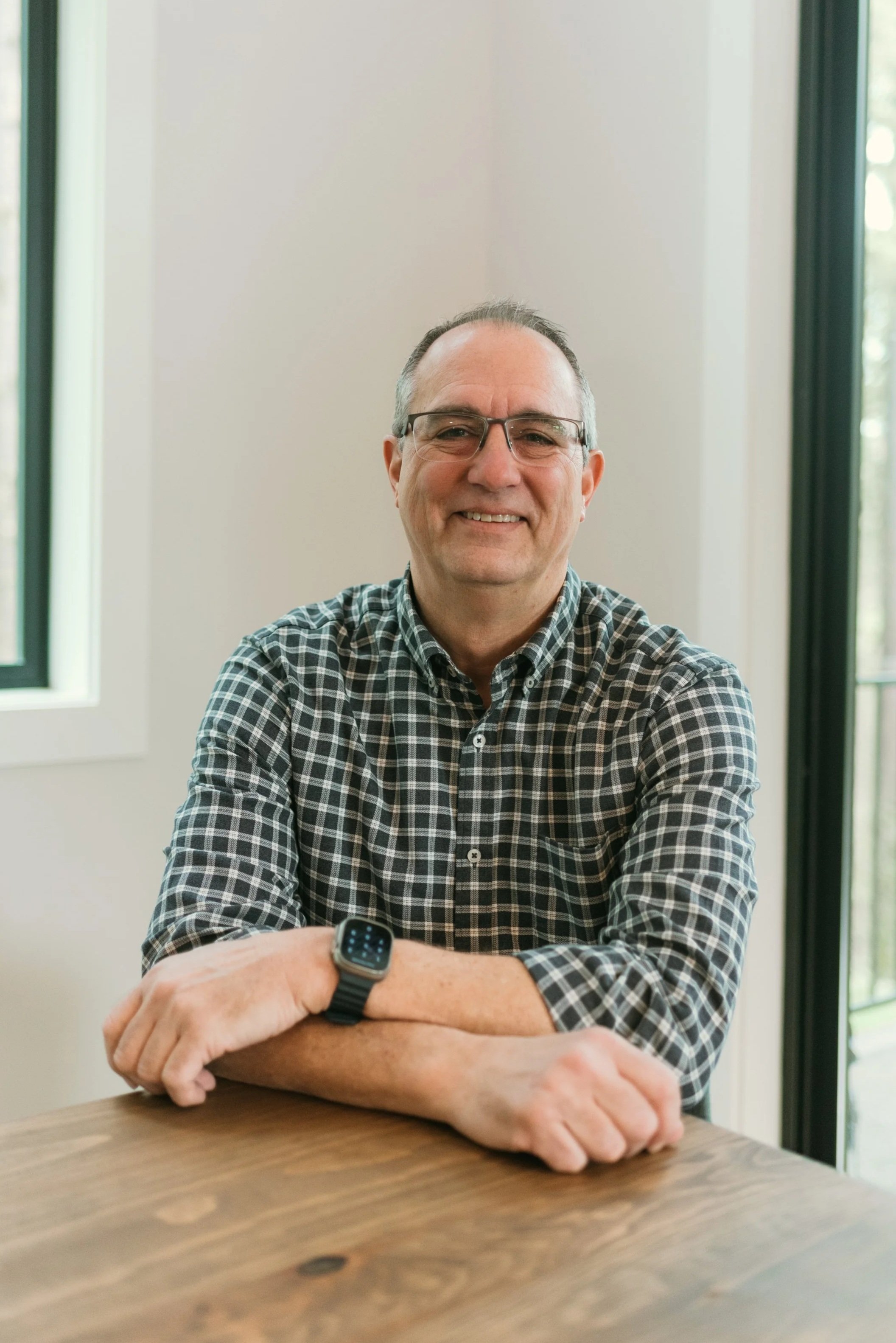 A middle-aged man with glasses and a checked shirt smiling while sitting at a wooden table, with large windows in the background and a smartwatch on his wrist.