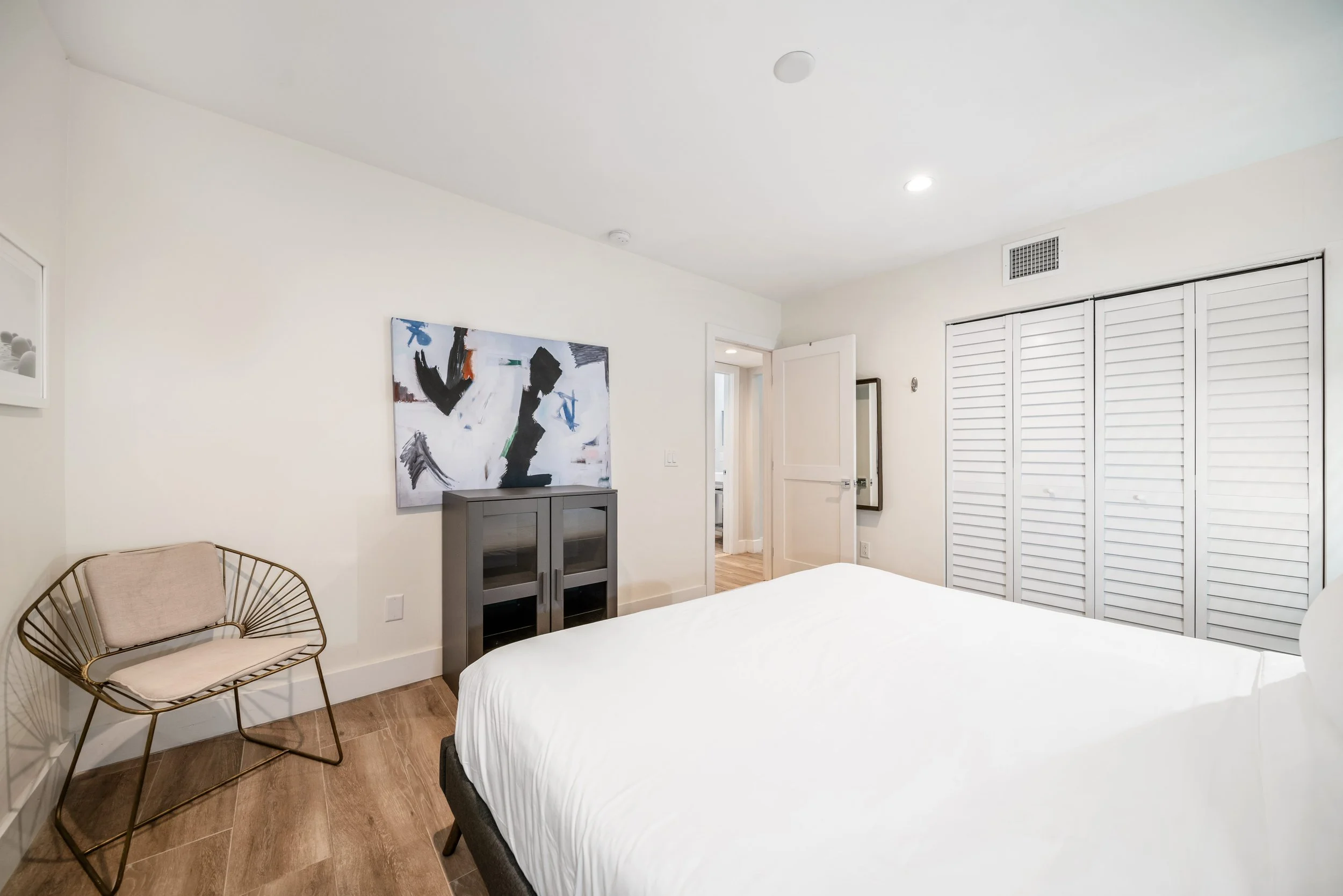 A bedroom with a bed, a gold chair with a beige cushion, a gray cabinet, a colorful abstract painting, a door, a mirror, a closet with white louvered doors, and wooden flooring.