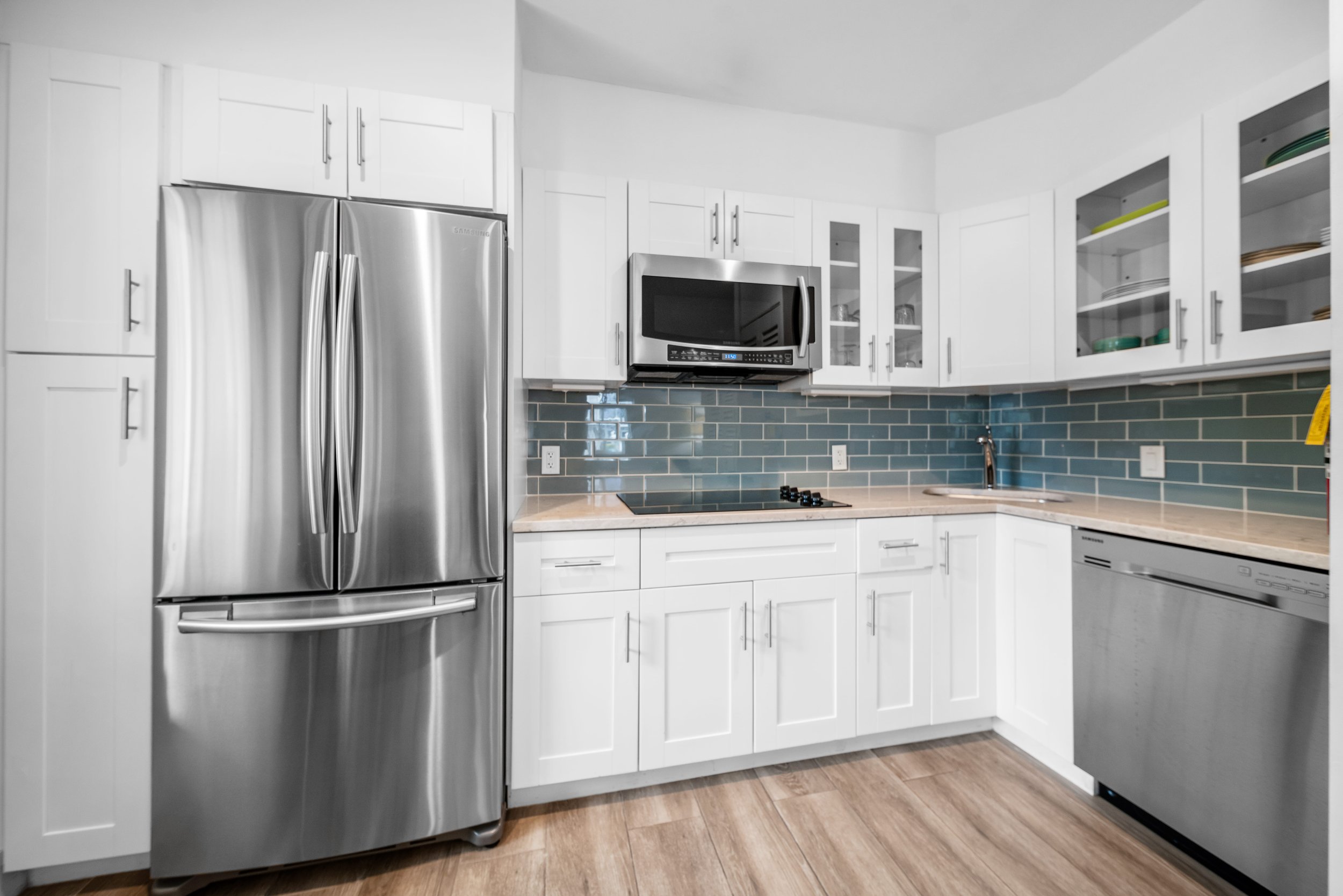 Modern kitchen with white cabinetry, stainless steel refrigerator and dishwasher, blue tile backsplash, microwave, black cooktop, and wooden floor.