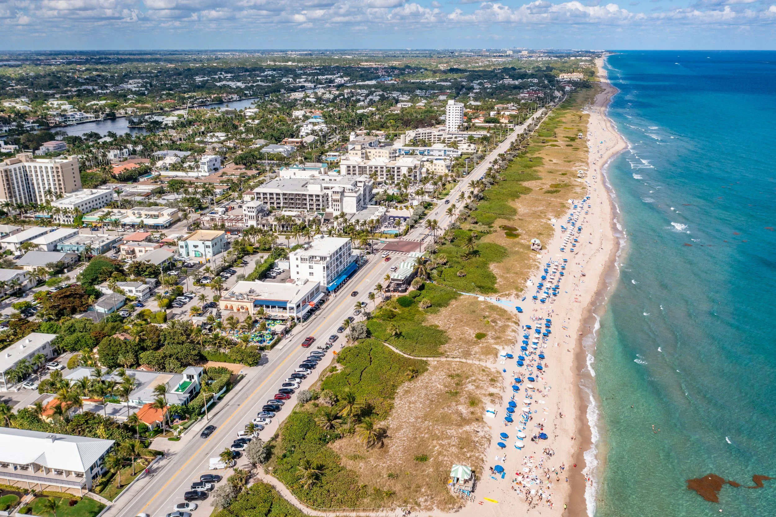 Aerial view of a beachfront town with a sandy beach, blue umbrellas, and a cityscape with buildings and streets