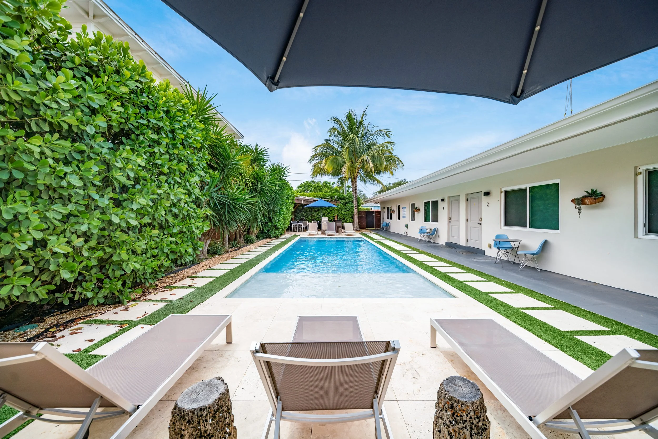 A backyard with a narrow swimming pool, green hedges, palm trees, and a white building with windows and doors, featuring outdoor seating and a large umbrella.