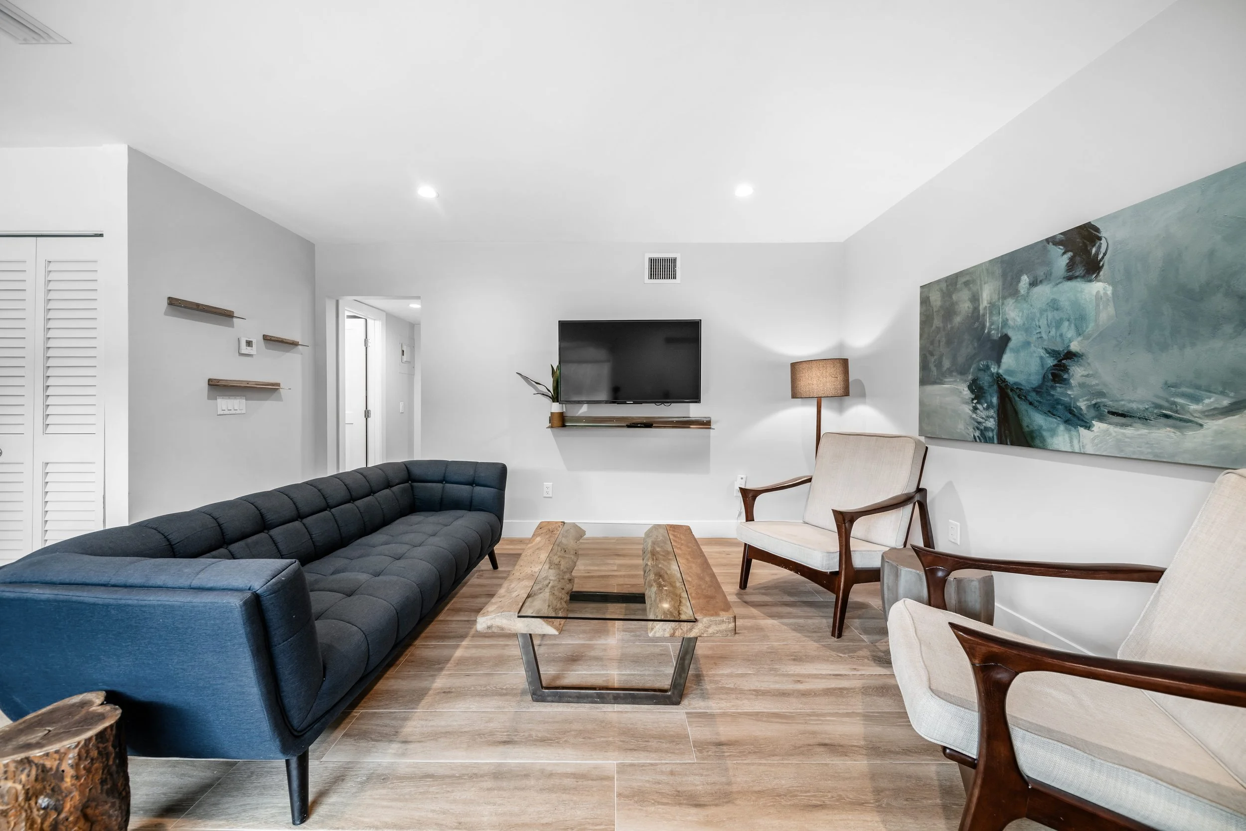 Modern living room with a dark blue sofa, two beige armchairs with wooden arms, a wooden coffee table with a glass top, wall-mounted flat-screen TV, large landscape painting, and a floor lamp.