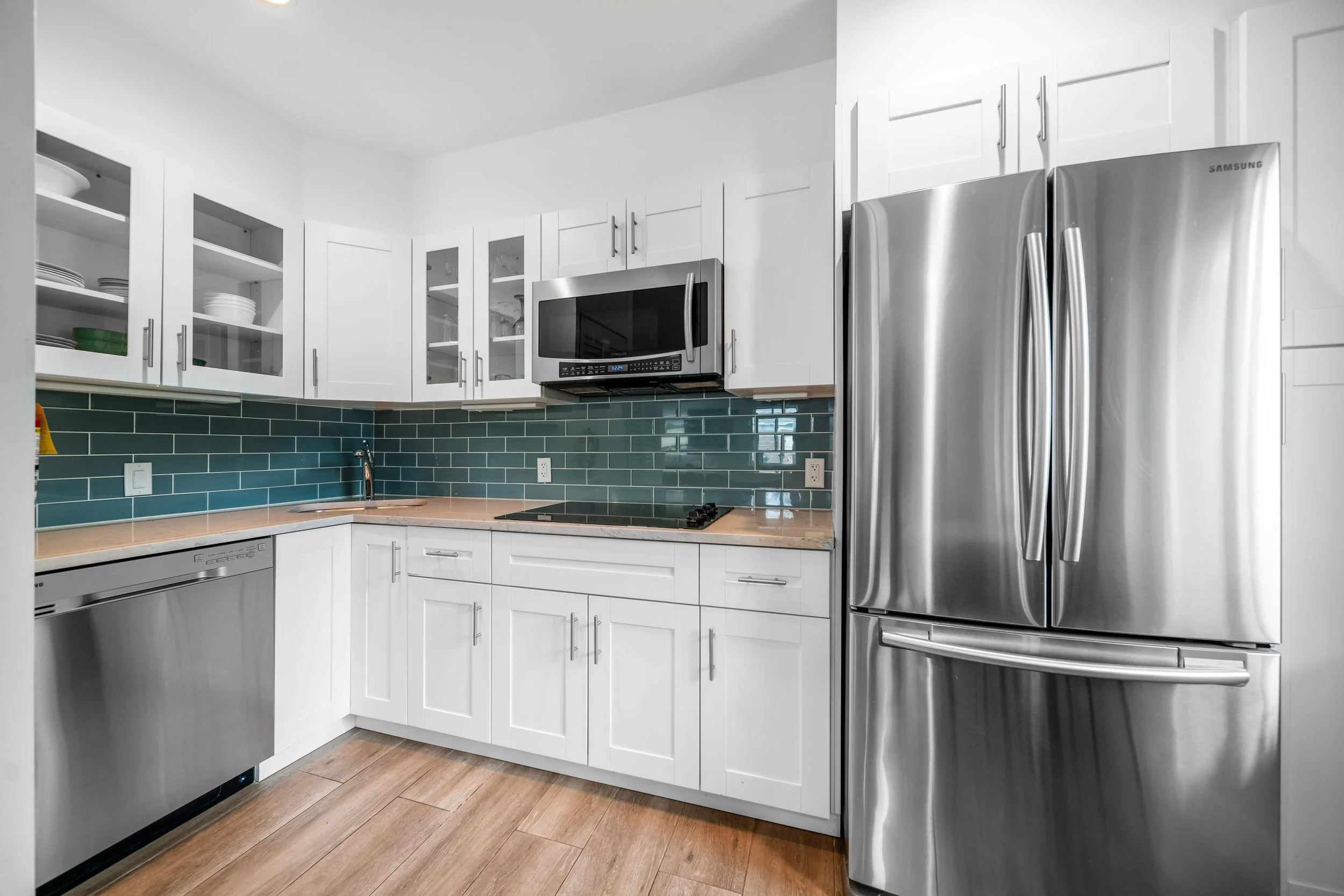 Modern kitchen with white cabinets, stainless steel appliances, green tiled backsplash, wood flooring, microwave, dishwasher, double-door refrigerator, and a cooktop.