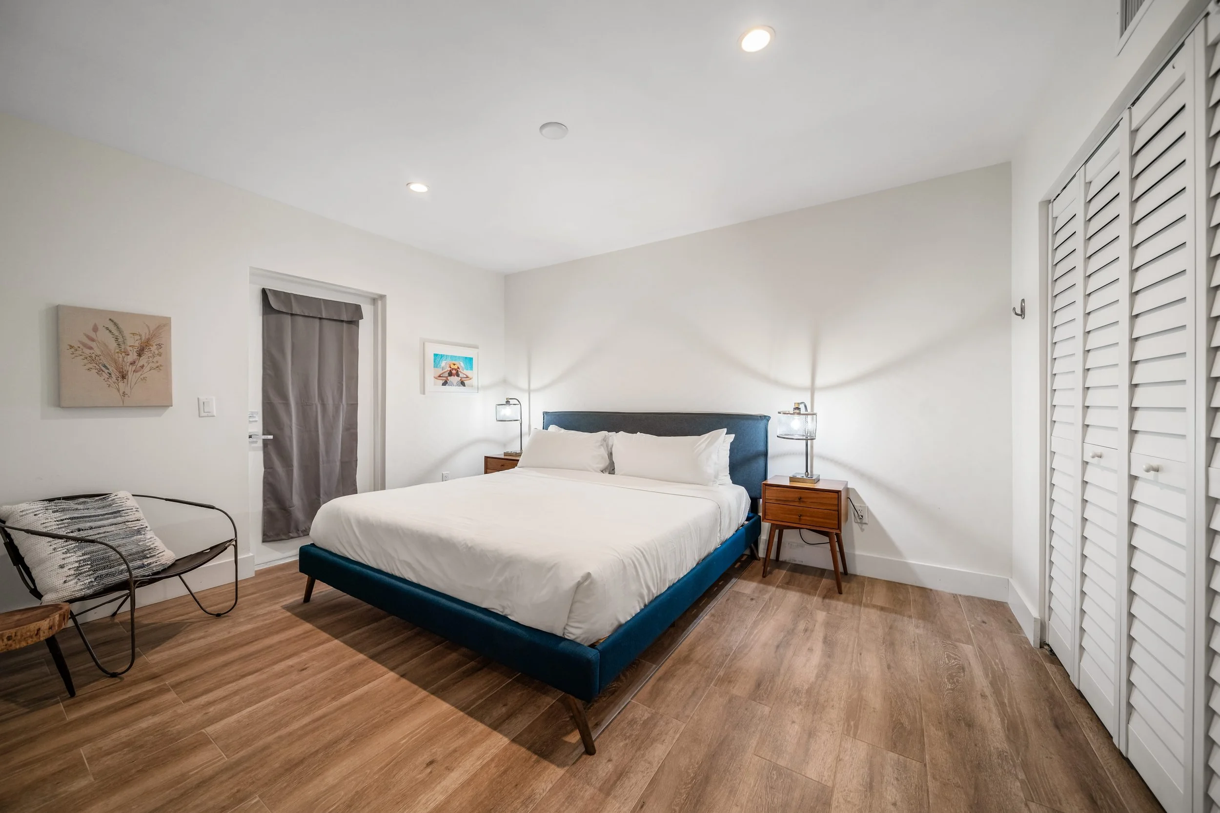 A bedroom with a King-sized bed with white bedding and a blue upholstered headboard, two wooden nightstands with table lamps, a chair with a cushion, and a closet with louvered doors.