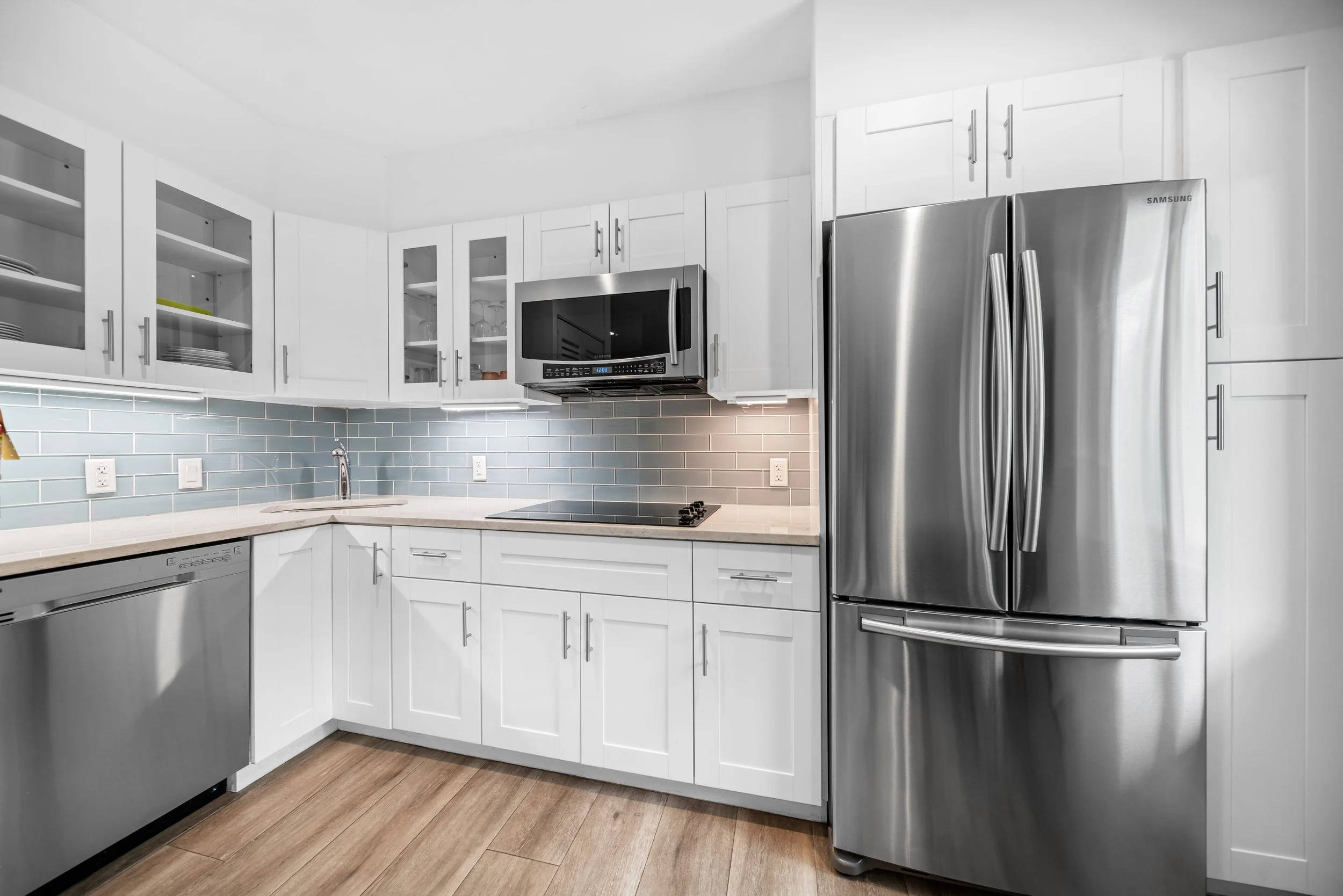 Modern kitchen with white cabinets, stainless steel refrigerator, microwave, dishwasher, blue tile backsplash, and wooden flooring.