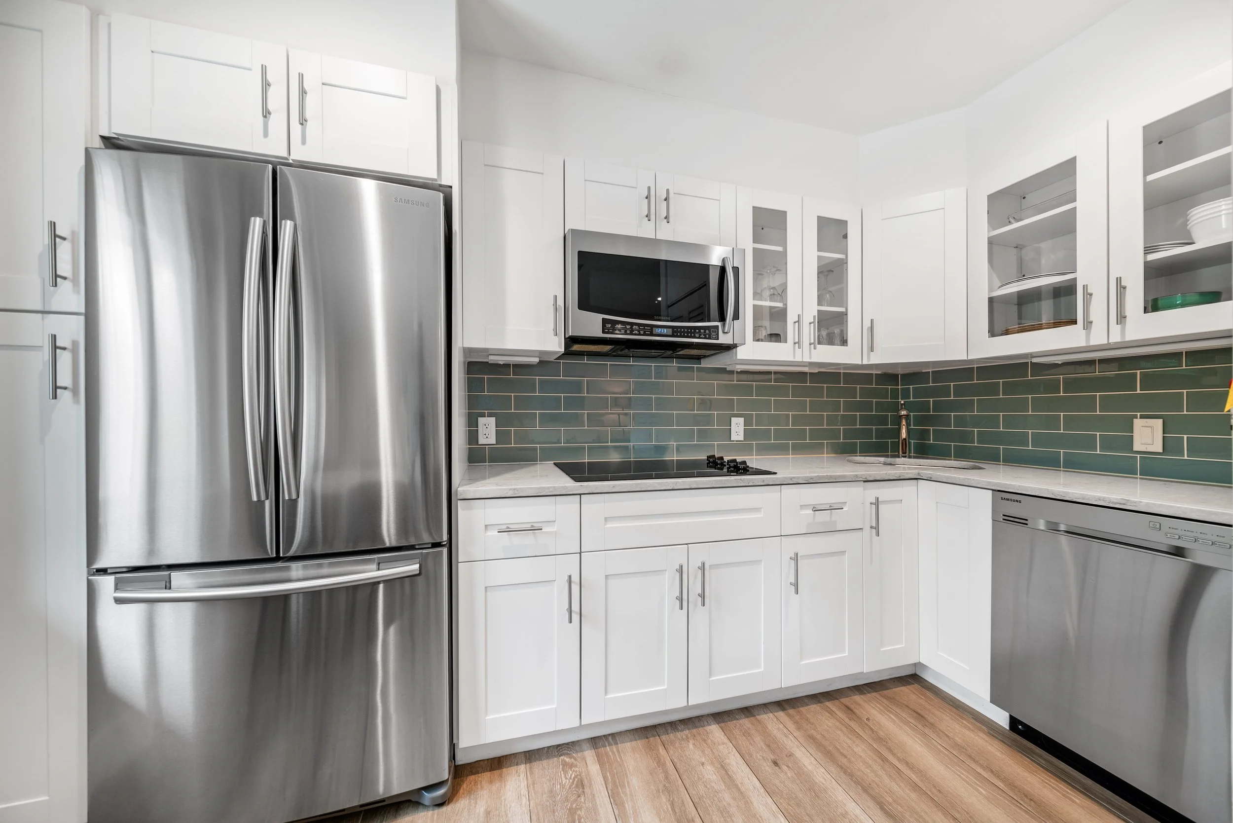 Modern kitchen with stainless steel refrigerator, microwave, dishwasher, white cabinets, green tile backsplash, wooden floor, and marble countertops.