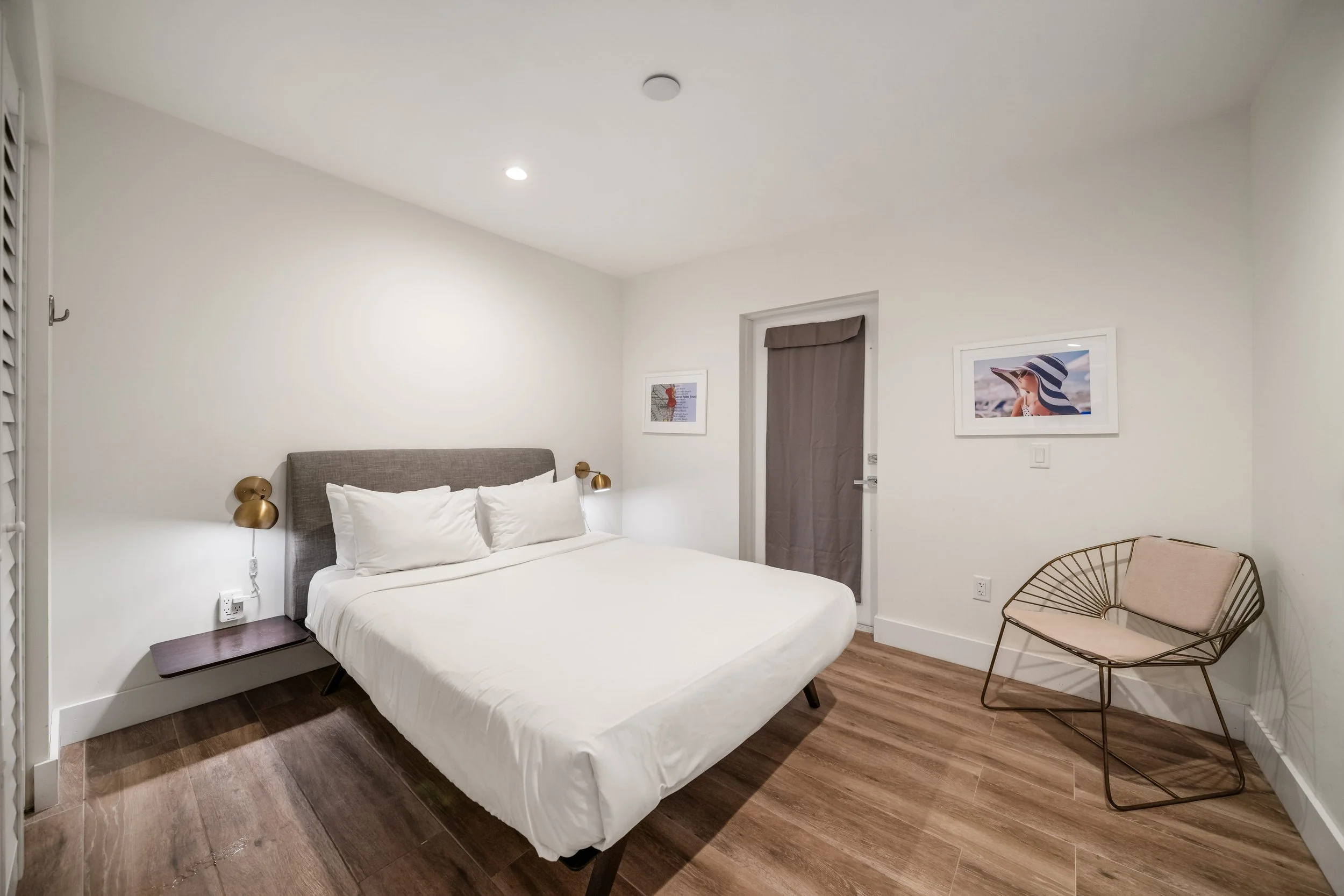 A minimalist bedroom with a bed, side table, wall-mounted lamps, two framed photographs on the wall, a chair, and a door with a curtain.