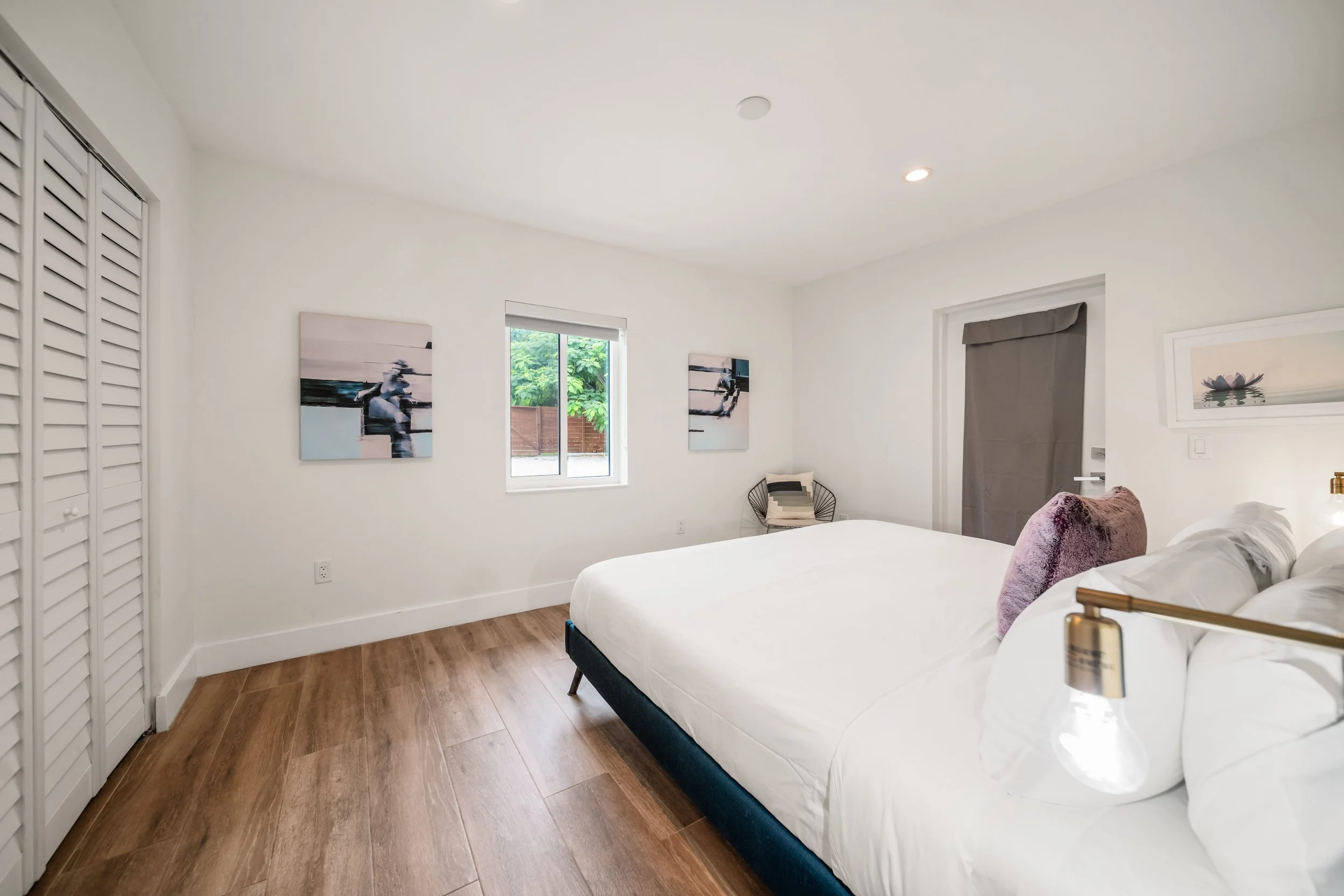 A modern bedroom with white walls, wooden flooring, a bed with white bedding and pink pillows, two artworks on the wall, a small seating area with a chair and throw pillows, a window showing greenery outside, and a door covered with a gray curtain.