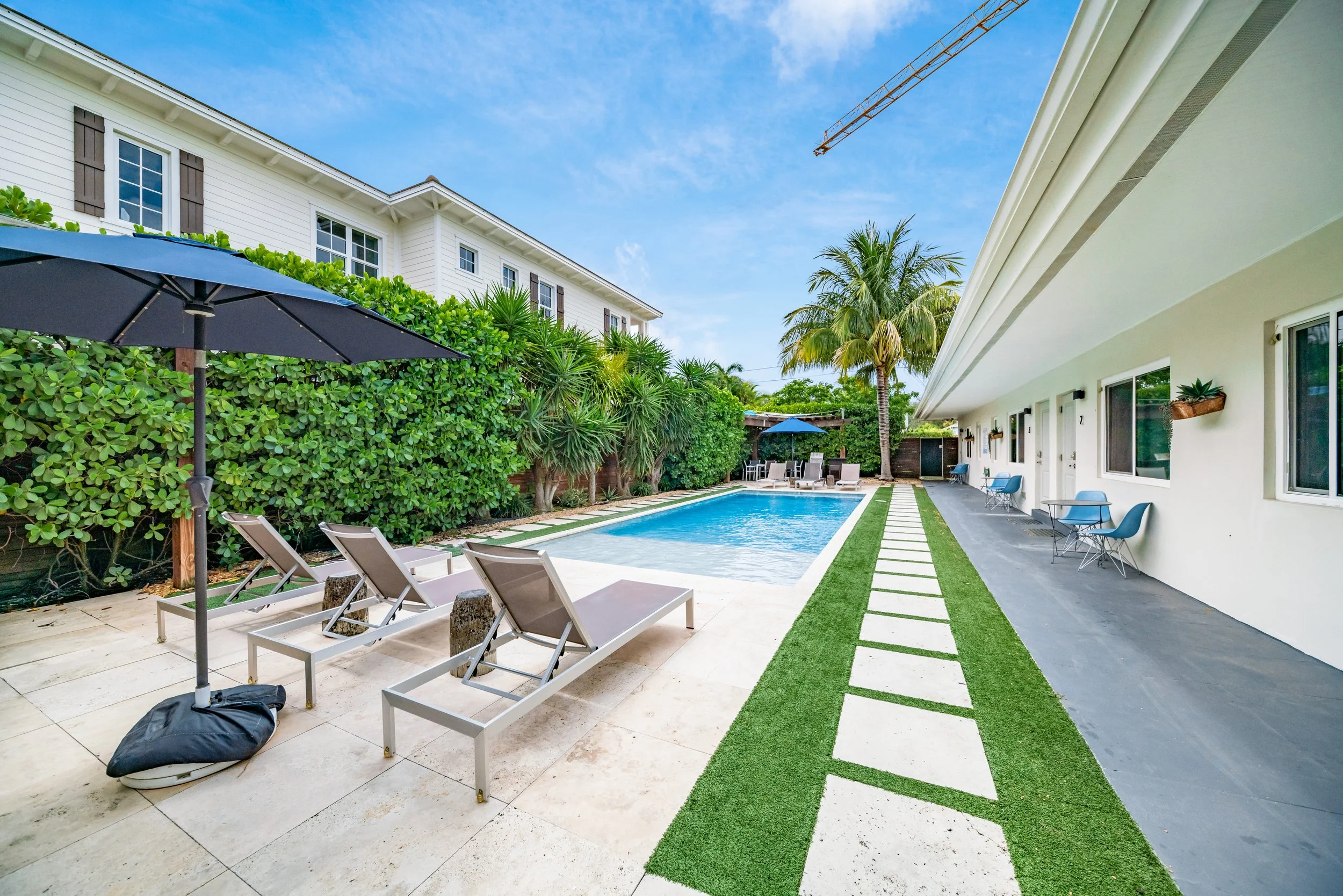 A backyard with a rectangular swimming pool surrounded by lounge chairs, a large umbrella, palm trees, and a white house with a patio and seating area.