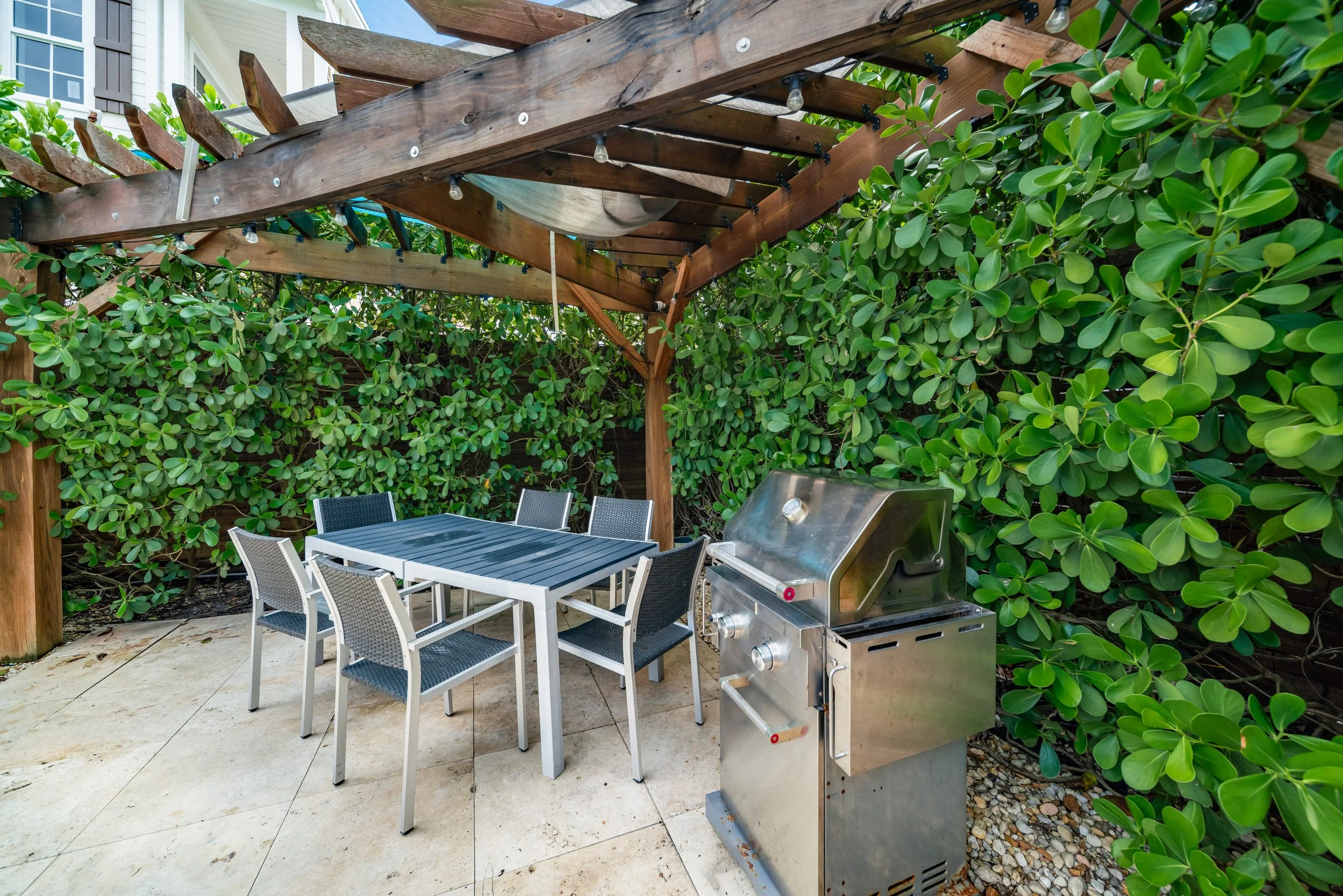 Outdoor patio with a black and silver dining table, six black chairs, a stainless steel grill, surrounded by lush green foliage, under a wooden pergola with string lights.