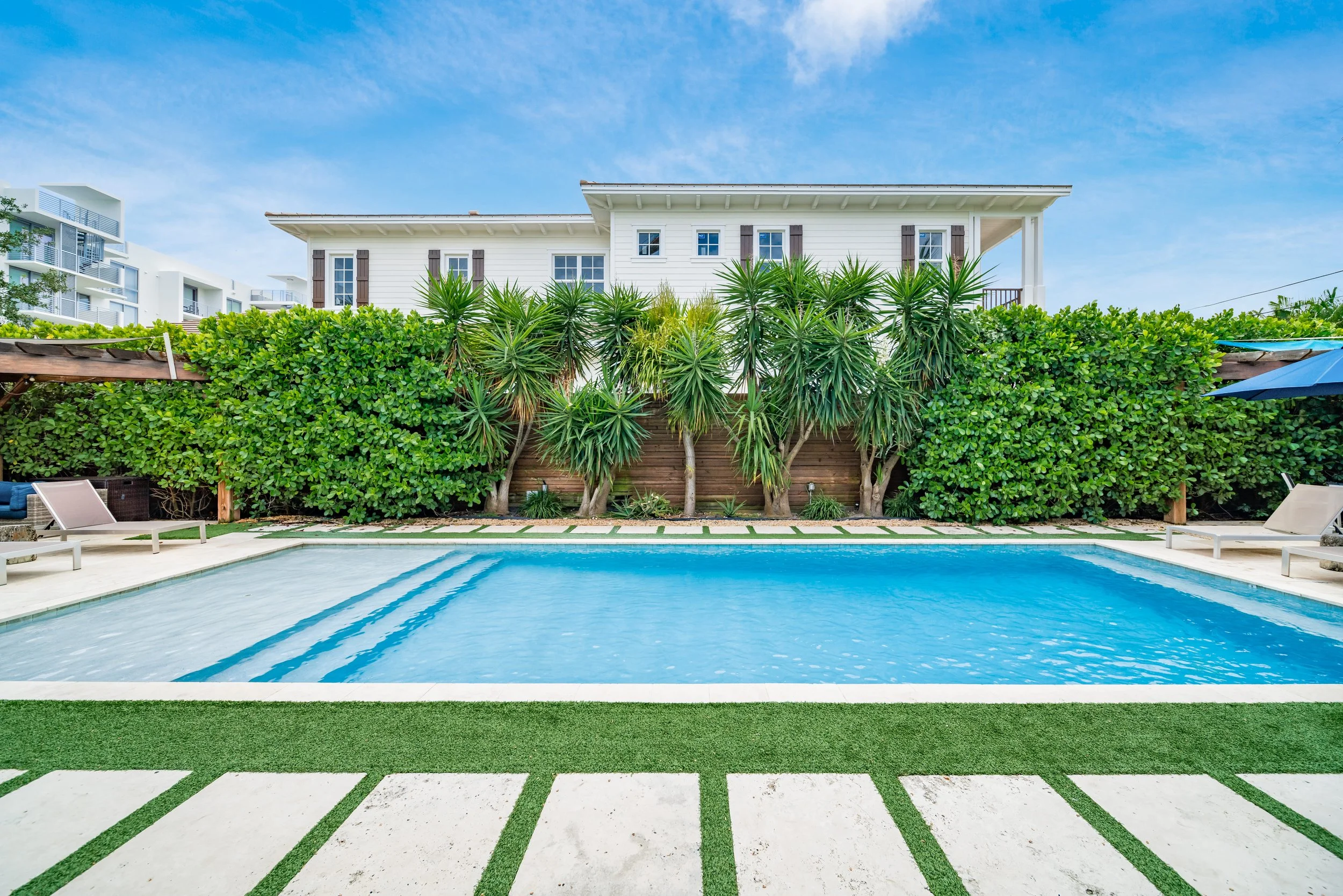 Swimming pool with lounge chairs, green bushes, and palm trees, with a white house in the background under a blue sky.