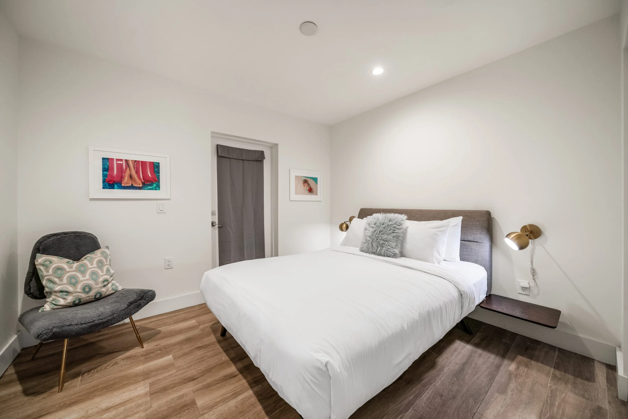 A modern bedroom with white walls and wooden floor, featuring a large bed with white bedding, gray headboard, and decorative pillows. There is a gray chair with a patterned pillow, two framed pictures on the wall, and wall-mounted brass reading lamps