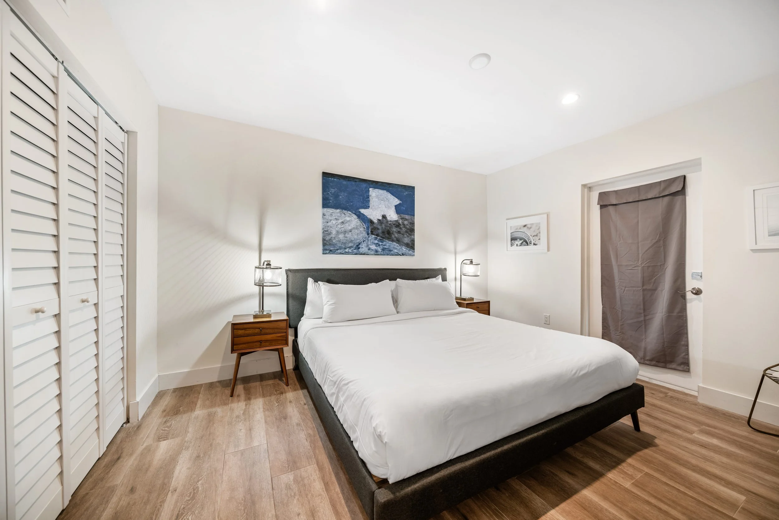 A modern bedroom with a large bed, white bedding, and wooden nightstands with lamps on either side. Wall art is displayed above the bed, and there is a closet with white louvered doors on the left. A window with a gray curtain is on the right.
